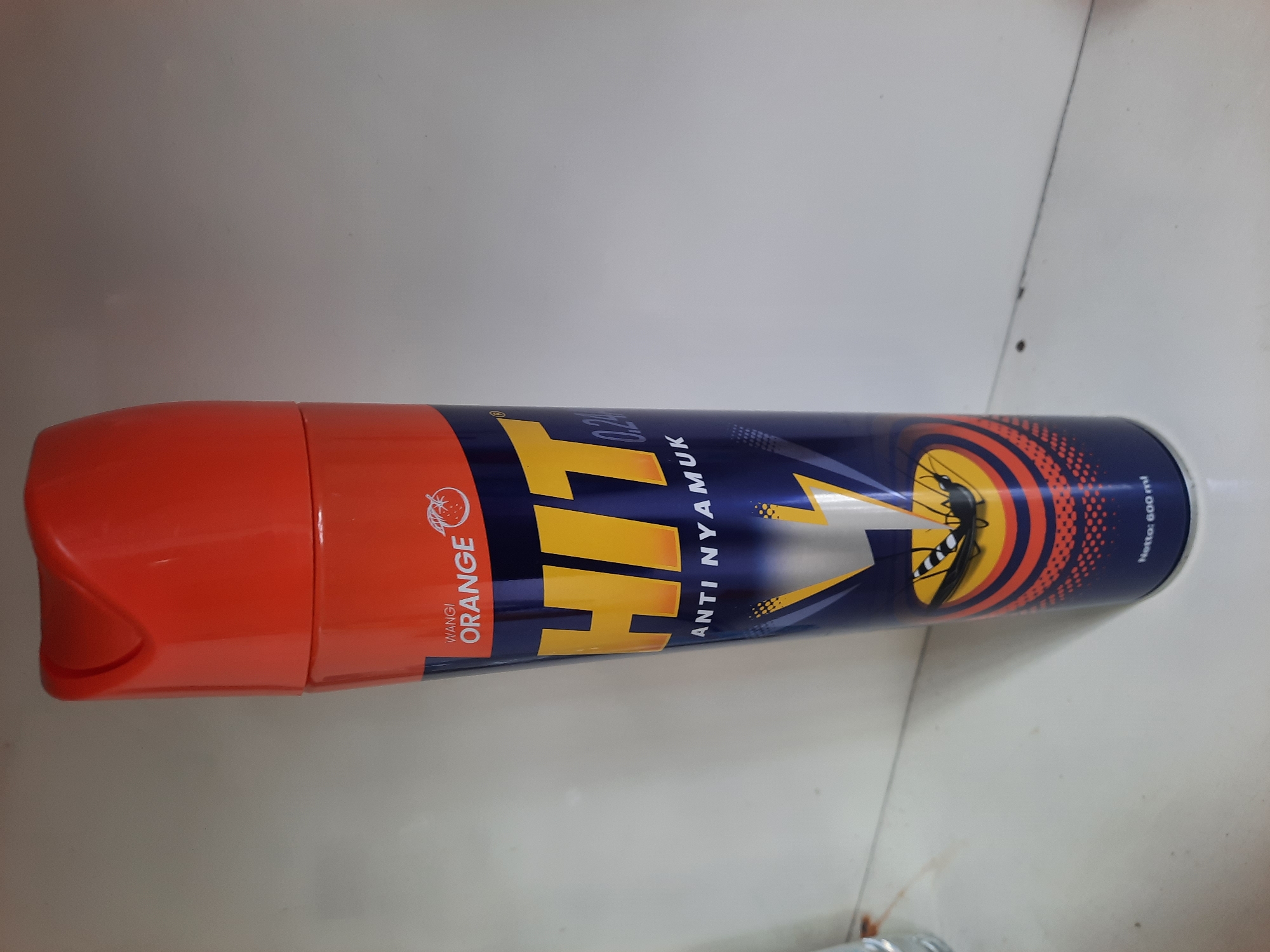 The image shows a can of "HIT" mosquito repellent with an orange scent. The can is primarily navy blue with a bright orange cap. The "HIT" logo is large and yellow, with the text "ANTI NYAMUK" (meaning "anti-mosquito") below it. There is an illustration of a mosquito being struck by lightning. At the bottom of the can, the text "Netto: 500 ml" is visible. The can is standing upright on a white, flat surface, likely a shelf or countertop, with a light gray wall in the background. There are no people or specific landmarks of Lhokseumawe, Indonesia visible in the image; it is a general product shot.