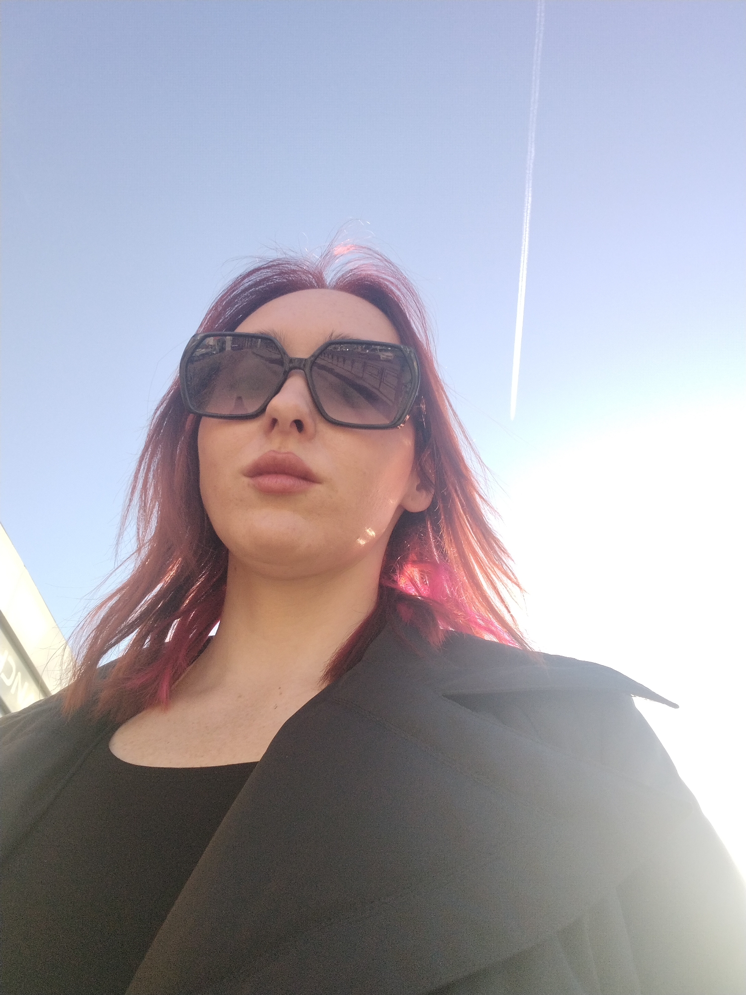 A low-angle shot features a woman with dark red hair and large, square sunglasses. She is wearing a black top and a black coat or jacket. The background is a clear, light blue sky with a visible contrail from an airplane. The sun is shining brightly from the right, causing lens flare and overexposure in that area of the image. A building with signage can be seen partially in the lower left corner. Based on the architectural style and signage, the location is likely Izhevsk, Russia.