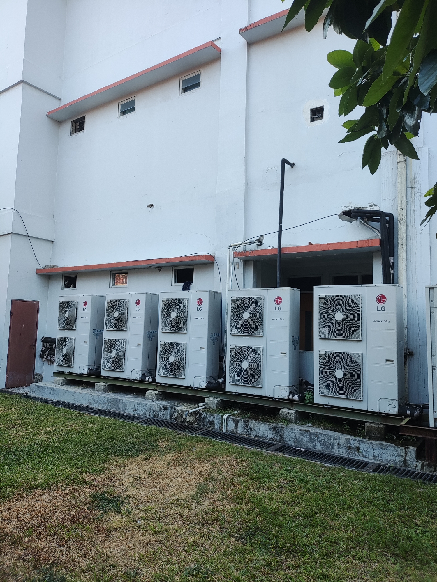 An exterior, eye-level, wide shot captures a wall of an institutional building in Cirebon, Indonesia, with a row of outdoor air conditioning units situated along its base. The wall is painted a pale white, with a prominent orange-red trim running horizontally above the air conditioning units. Several small, rectangular windows are visible along the upper portion of the wall, some with dark grates. To the left, a dark brown, single door is present.
Below the wall, a concrete platform supports a line of six LG brand air conditioning units, each appearing to be a multi-split system, indicated by the "Multi V" branding. These units are white and feature circular vents with a dark grille. They are arranged in two columns, with three units in the left column and three in the right. Piping and electrical conduits are visible connecting to the units and running along the wall.
In front of the platform, a section of green grass extends to the foreground, with some areas showing signs of dryness or browning. A metal grate drain runs along the edge of the concrete platform, separating it from the grassy area. A mature tree with large green leaves partially obscures the upper right corner of the image, casting shadows on the building. The lighting suggests it is daytime, possibly late afternoon given the angle of the light. There are no people visible in the image.