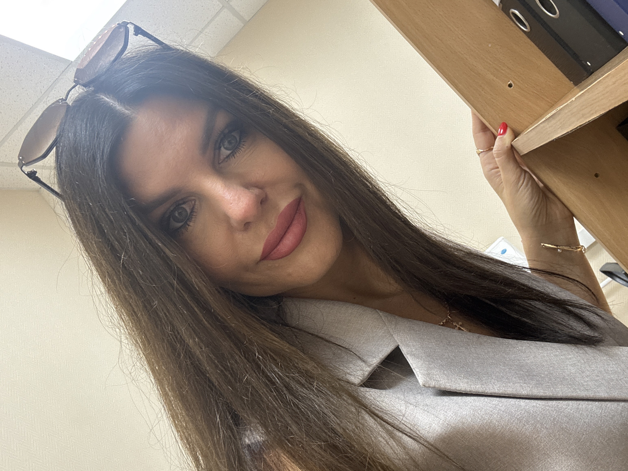 In this high-angle, close-up shot taken indoors, a woman with long, dark brown hair and makeup, including pink lipstick and eyeliner, is pictured. She is wearing a light gray blazer. Sunglasses are perched on top of her head. Her right hand, with red nail polish, is resting on a wooden shelf. The background appears to be an office setting, with a white ceiling with fluorescent lighting and a beige wall. The image does not provide any visual cues to suggest a specific location within Chaikovsky, Russia.