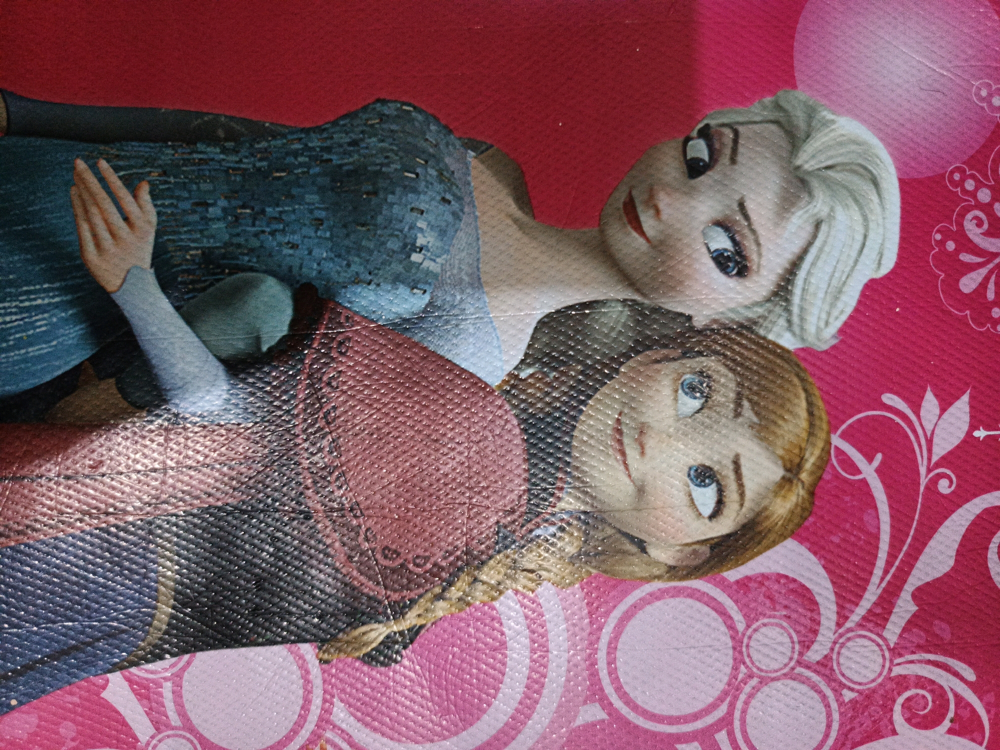 The image shows a close-up, overhead view of a patterned mat or fabric featuring cartoon characters resembling Elsa and Anna from the movie Frozen. The characters are depicted in a stylized, doll-like manner, with large eyes and soft features. Elsa, on the left, is shown with blonde hair and a blue dress. Anna, on the right, has auburn hair and a red dress. They are positioned in a way that suggests they are interacting or posing together. The background is a vibrant pink with a textured, grid-like pattern, overlaid with white floral and circular designs. The lighting is bright, highlighting the colors and textures of the material. The image appears to be taken in a domestic setting, likely within Pabelan, Indonesia, given the context.