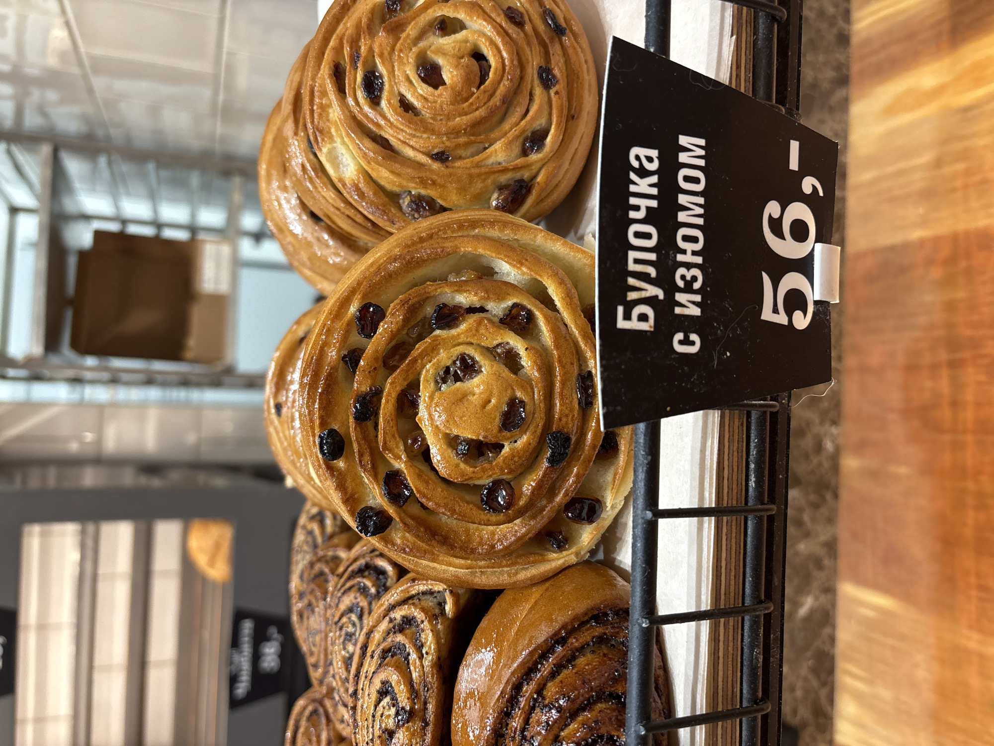 The image shows a selection of pastries displayed in a black wire basket. The pastries include raisin buns and poppy seed rolls. A black sign with white text reading "Булочка с изюмом 56,-" (Bun with raisins 56,-) is placed next to the buns. The location is Kudrovo, Russia, based on the Cyrillic writing and the potential availability of such baked goods in the region. The scene appears to be inside a bakery or cafe.