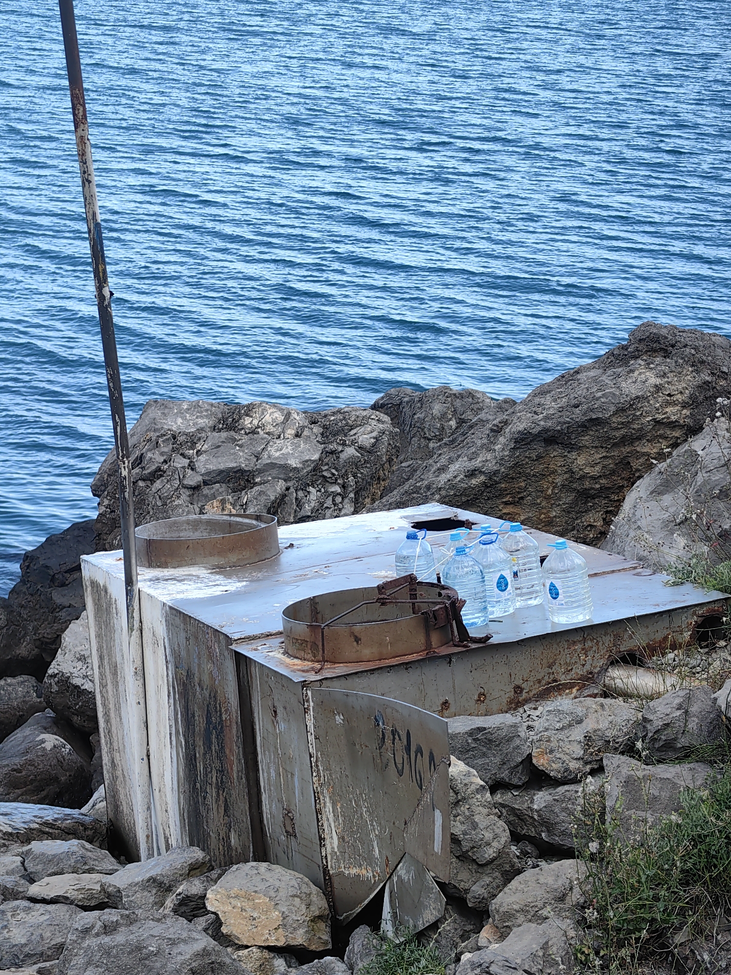In Chaika, Ukraine, several large plastic water bottles with blue caps are arranged on a metallic surface atop a large, weathered, metal structure situated amongst rocky terrain. A rusty metal pole is affixed to the left side of the structure, extending upwards and partially obscuring the bright blue, rippling water of the sea in the background. The metal structure appears aged, with visible rust and discoloration. The surrounding environment is composed of large, grey rocks, some covered in sparse green vegetation. The scene appears to be outdoors, likely near a coastline.