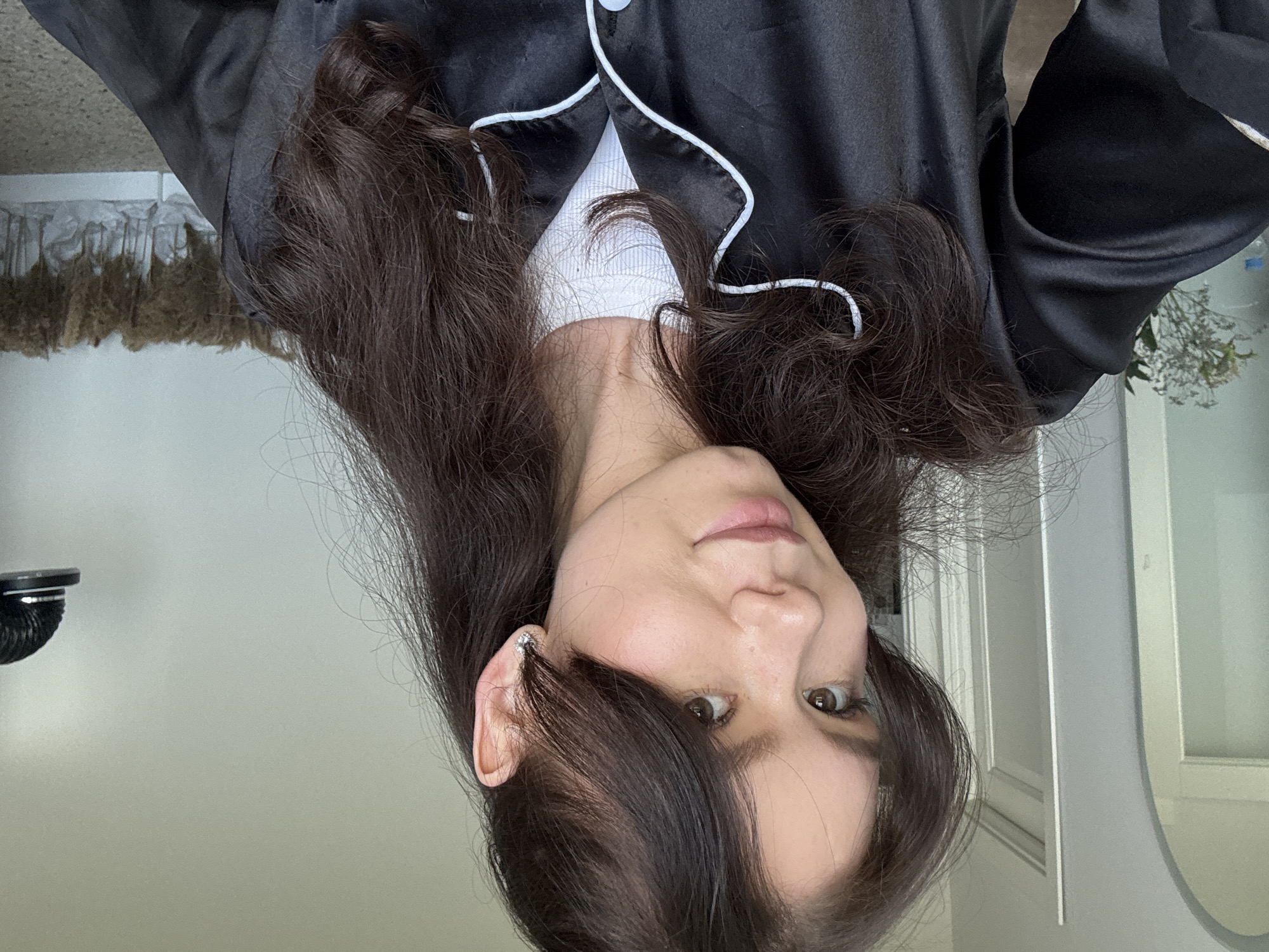 A person with dark hair and fair skin is facing the camera while wearing a black pajama top with white piping. The person has a silver earring in their left ear. The background includes a white wall, a round mirror, and a glimpse of an object with brown hanging fibers and possibly some flowers near a window. The location appears to be indoors.
