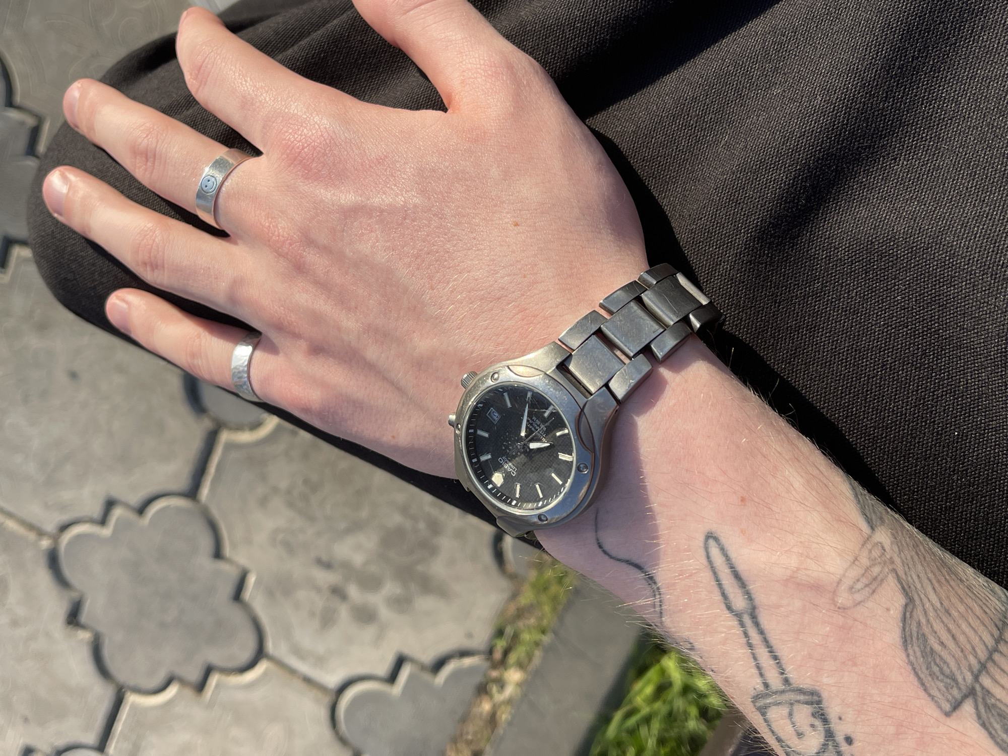 A close-up shot captures a person's left hand and forearm. The hand features two silver rings, one on the index finger with a smiley face and another on the pinky finger. A silver wristwatch with a dark face is worn on the wrist. The forearm has visible tattoos. The hand rests on a dark brown fabric. The background shows a patterned stone pavement and some green foliage. The image appears to be taken outdoors in daylight. No specific location within Krasnoyarsk, Russia, can be ascertained from this image alone.