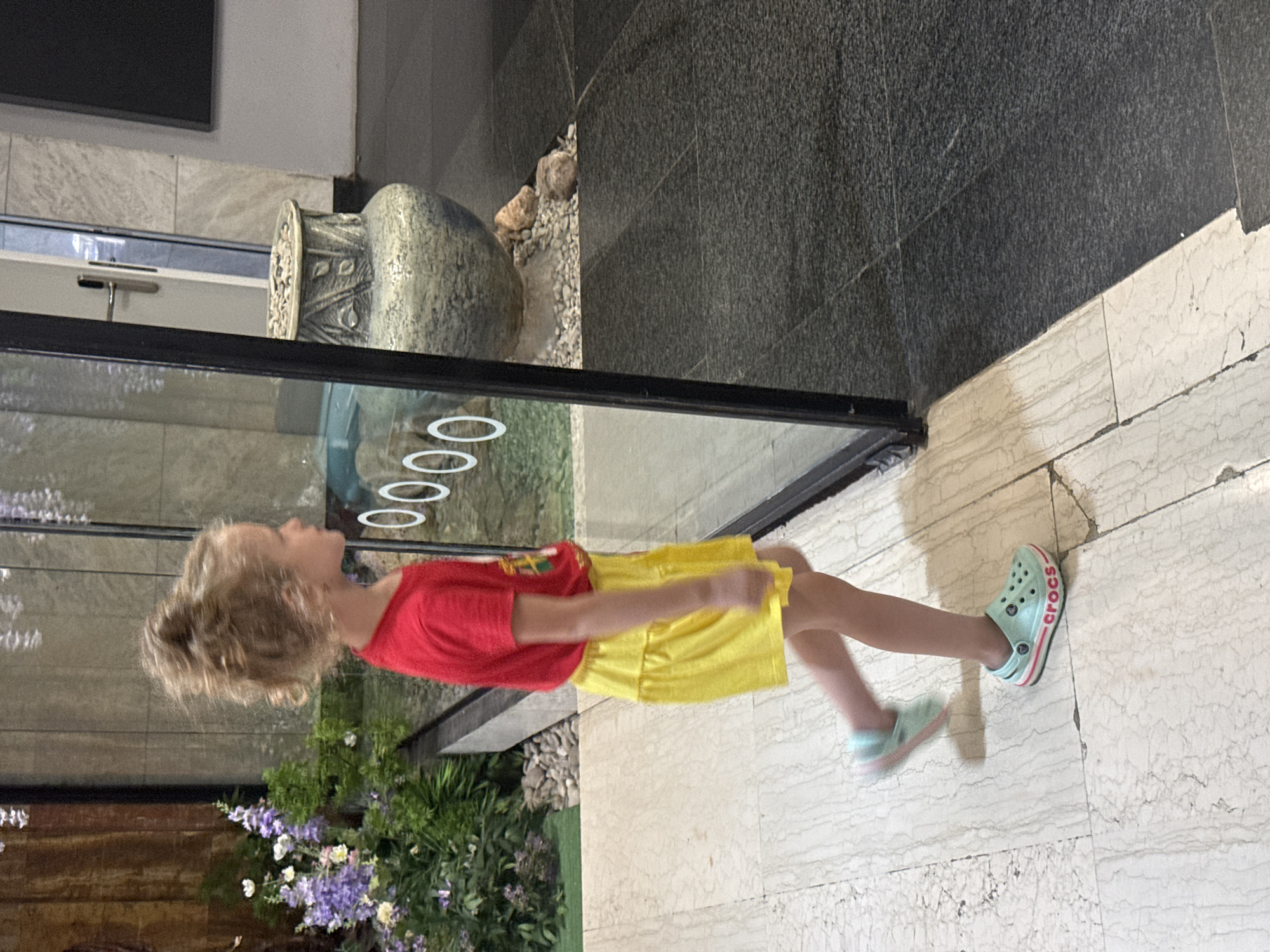 A young girl with blonde, curly hair tied up in a bun, wearing a red t-shirt and a yellow skirt, walks past a glass partition. She is wearing light blue Crocs with pink and white accents. In the background, through the glass, a large, decorative pot and some potted plants with purple and white flowers are visible. The floor is tiled with a light-colored marble pattern on the left and dark gray tiles on the right. The scene appears to be an indoor walkway or public space, possibly a hotel or a shopping center, in the city of Yalta, Ukraine.