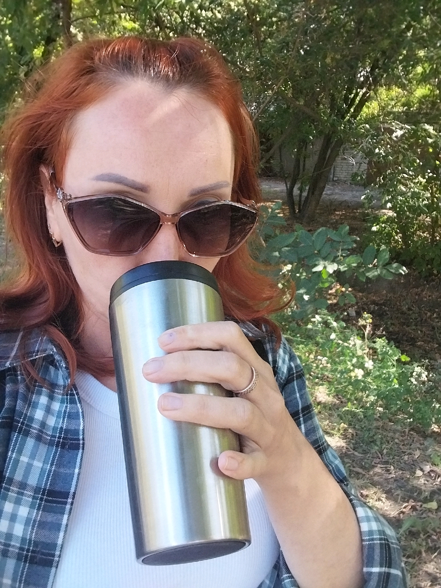 A woman with auburn hair and sunglasses is holding a silver thermos cup to her lips, as if drinking. She is wearing a white ribbed t-shirt and a blue and grey plaid shirt. A ring is visible on her right hand. The background is out of focus and appears to be a park or wooded area with trees and foliage. The lighting suggests it is daytime. The image is a close-up shot, focusing on the woman's face and upper body.