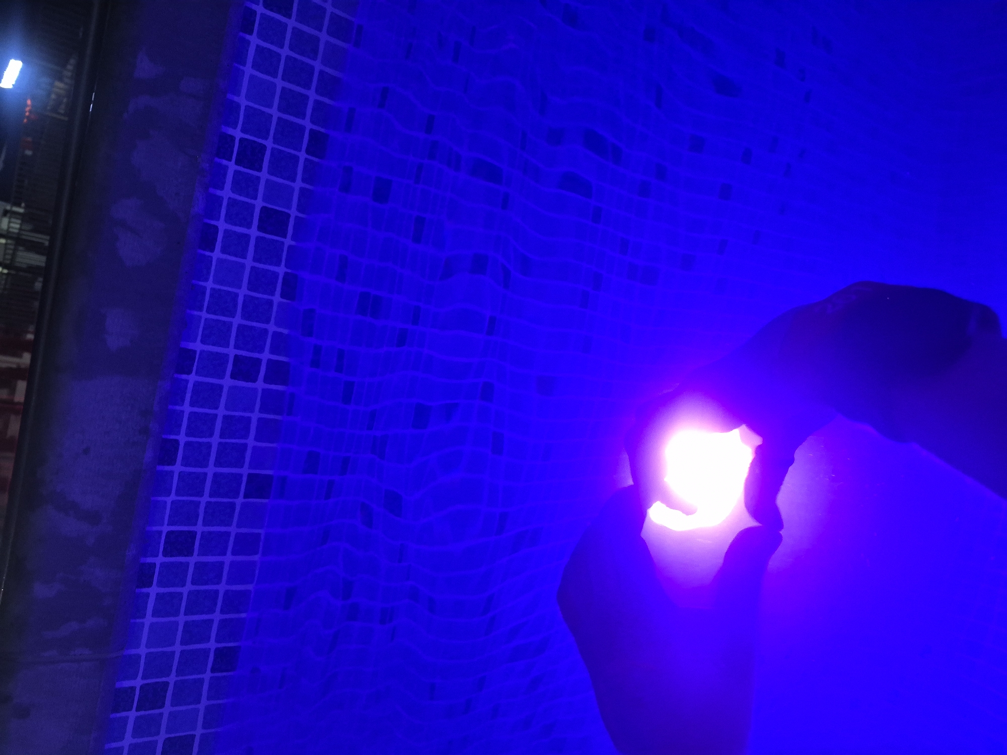 The image captures a vibrant and intimate scene, likely at a swimming pool in Votkinsk, Russia. The primary subjects are a pair of hands forming a heart shape around a powerful, bright white light source. These hands are positioned in the lower right portion of the frame, largely silhouetted against the intense glow.
The activity taking place involves a person holding a light underwater or just above the surface, with their hands deliberately arranged to create a heart shape. The surrounding water is illuminated by an artificial deep blue or violet light, creating a dramatic and monochromatic atmosphere.
Notable details include the textured surface of the pool's wall or floor, visible as a grid of small, square tiles along the left and central parts of the image. The water surface shows wavy distortions, reflecting the light and possibly movement. In the upper left background, beyond the tiled edge, a brighter white light fixture is faintly visible, suggesting an indoor pool or nighttime setting. The bright central light within the heart shape dominates the right side of the image, contrasting sharply with the deep blue environment. No visible text is present.