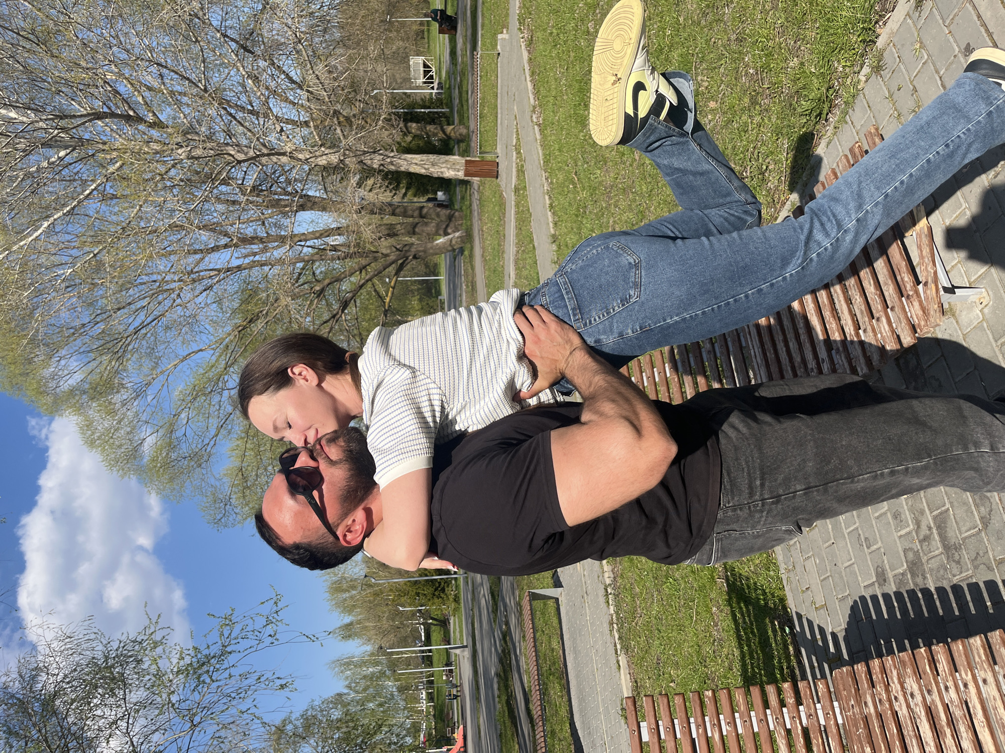 In Ulyanovsk, Russia, a man and woman are on a park bench. The woman, wearing jeans and a striped shirt, leans back on the man, who wears a black t-shirt and jeans. They are kissing. The man sits on the bench while the woman is held by him. The area includes grass, trees and paved paths.