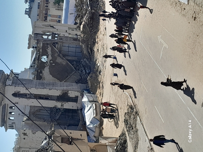 The image captures a wide-angle view of an urban landscape in Khan Yunis, Palestinian Territory, characterized by significant structural damage under a clear sky. In the background, a large, multi-story building, likely a mosque given its architectural features including domes and arched windows, stands severely damaged, with large sections of its facade collapsed, exposing internal structures and leaving substantial piles of rubble at its base. Adjoining structures to the immediate left are also heavily damaged, contributing to the debris field.
In the midground and foreground, an asphalt road with visible white lane markings, including a double solid line, a single broken line, and a directional arrow, extends through the scene. The road edges are lined with sand and dirt, intermingled with smaller debris piles. Numerous people, estimated to be over 40 individuals, are present and in motion. On the left side of the road, a man in a dark jacket walks rightward. Further back on the left, a small group of 3-5 individuals stands near a bicycle amidst rubble.
Centrally, a woman in a dark abaya walks on the road carrying a large, flat, circular object with both hands. To the right of the road, a dense cluster of approximately 20-30 individuals, comprising adults and children, is gathered on the sandy ground, some holding bags or various items, suggesting a congregation or queue. Several other individuals, primarily women in dark attire and children, are scattered across the scene, walking on both the road and dirt paths. Strong shadows are cast by the people, indicating bright sunlight. Overhead utility wires are visible in the upper left portion of the image.