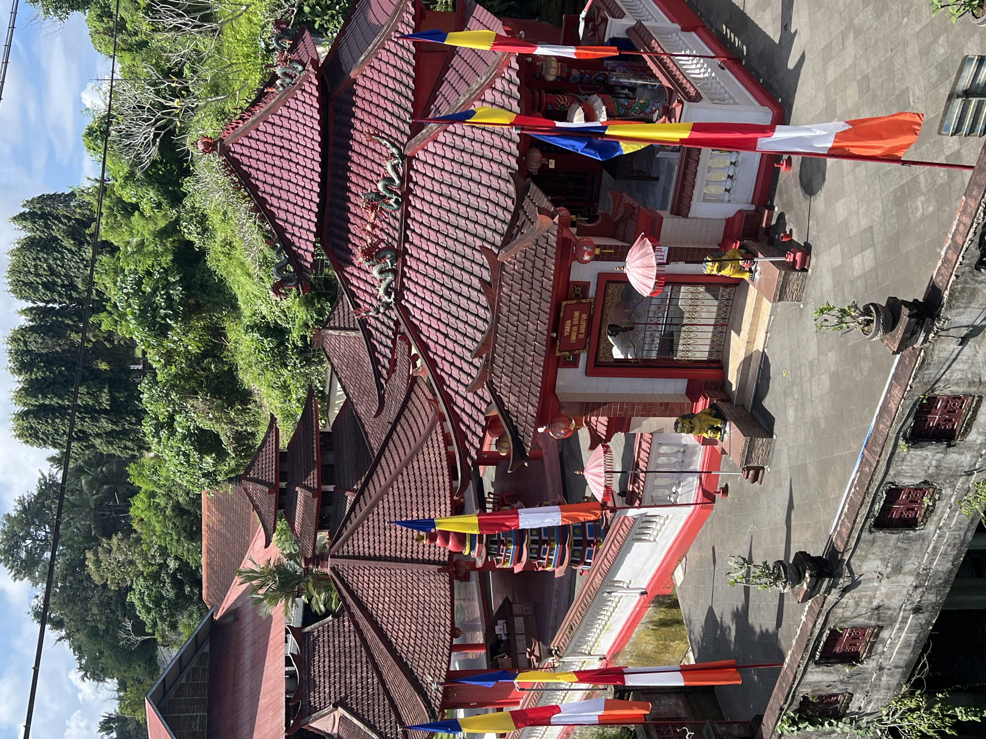 The image depicts a temple complex in Sukawati, Indonesia. The architecture features tiered red tile roofs adorned with dragon sculptures. The buildings are painted white with red trim and are decorated with colorful Buddhist flags. There are potted plants and religious objects like umbrellas and lanterns visible. A person is visible inside a temple building. The surrounding environment consists of dense tropical vegetation. The scene appears to be photographed from an elevated position.