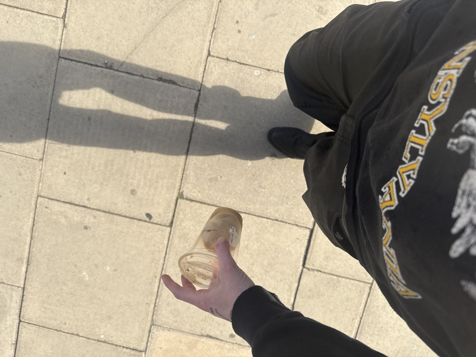 An overhead, first-person perspective photograph depicts a person standing or walking on a paved outdoor surface, likely a sidewalk, in London, United Kingdom. The ground is composed of light-colored, rectangular flagstones or tiles. Bright sunlight casts a prominent, elongated shadow of the person's lower body and legs onto the pavement. The person's left hand, which has a small dark tattoo on the wrist area, holds a clear plastic or glass cup. The cup contains a small amount of light brown liquid or residue at the bottom. A white rectangular label with "PROJECT X" in black text is affixed to the side of the cup. The person is wearing a black long-sleeved garment on the left arm, a black t-shirt with a yellow/orange graphic featuring partial text "...YLVANIA" and a dark graphic design beneath it, black pants, and a black shoe on the right foot.