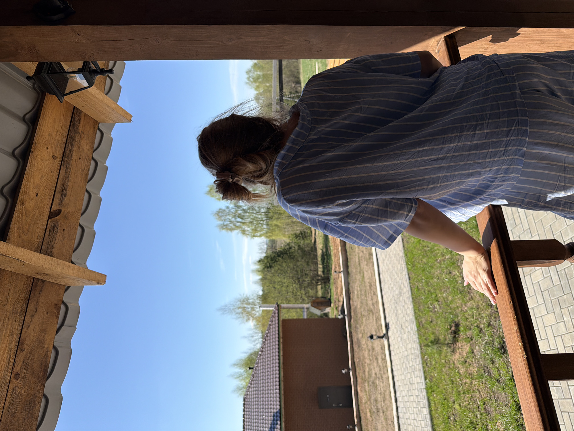 A woman stands on a wooden structure, possibly a porch or balcony, with a view of a landscape. The scene appears to be located in Kamennoye, Russia, based on the metadata. The woman, wearing a striped blue and white shirt, leans against the railing. She has her hair pulled back. The view includes a building with a dark roof, a walkway, grass, and trees under a bright blue sky. The wooden structure has visible beams and a light fixture.