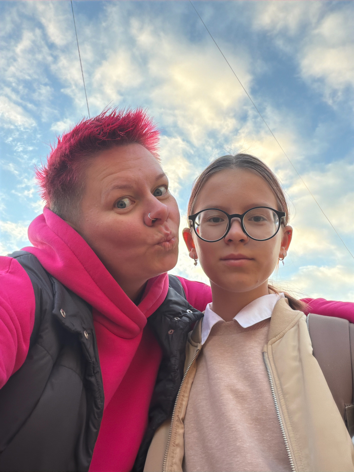 The image shows a selfie of two people, likely a mother and daughter, outdoors with a sky in the background. The person on the left has bright pink, spiky hair and is wearing a pink hooded jacket and a black vest. They are making a kissing face. The person on the right is wearing glasses, a white collared shirt, a beige sweater, and a beige jacket. The environment appears to be a city, possibly Chaikovsky, Russia, given the telephone wires visible in the sky.
