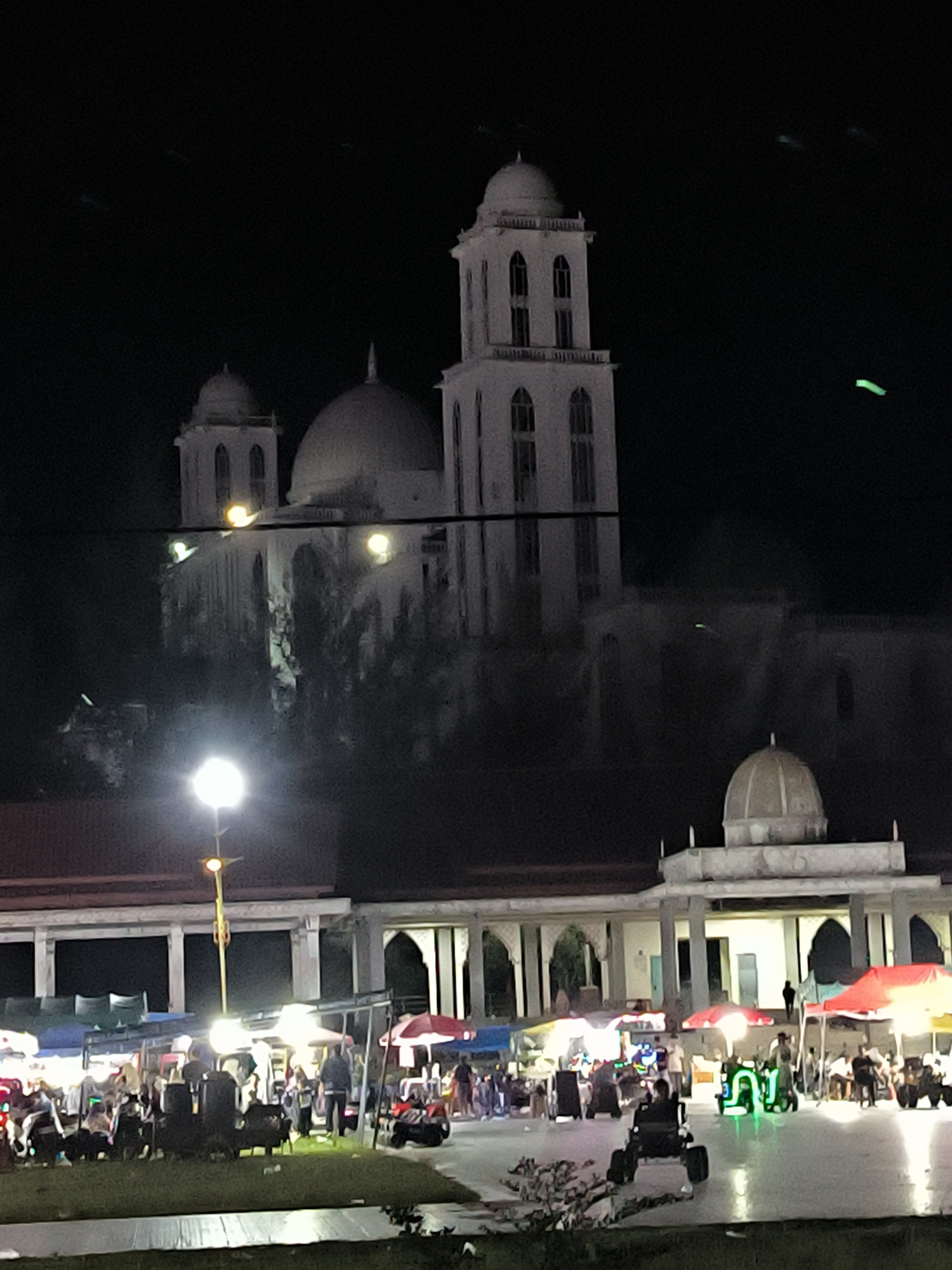 The image shows a night scene in Titibaro, Indonesia. In the background, a large, white building with a dome and a tower stands illuminated. In front of it is a lower building with arches and domes, under which a night market is taking place. People are gathered around various stalls with colorful umbrellas and lights. There are also small vehicles, possibly for children, with illuminated features. A street lamp illuminates the area.