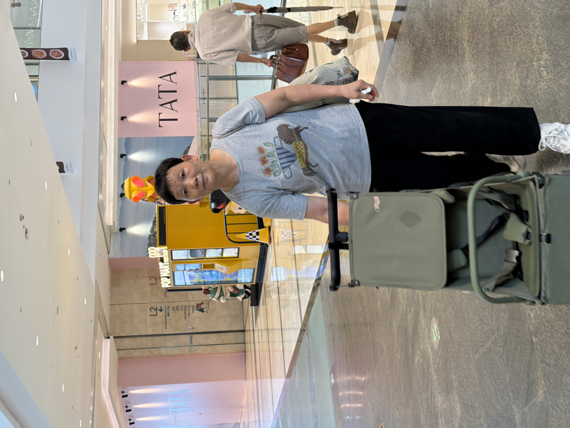 In a brightly lit indoor shopping mall located in Yuzhong District, China, a middle-aged East Asian woman is the central focus of the image. She is standing and looking directly at the camera, with her left hand near a light green stroller or cart positioned in front of her. She is wearing a grey t-shirt adorned with a graphic of a horse and flowers in a pot, and loose black pants. Her right hand is partially visible holding a patterned tote bag. In the background, another person, an East Asian male, is walking away from the camera, carrying a brown leather bag and an umbrella. The environment features polished floors, soft pink and beige walls, and illuminated displays, suggesting a modern retail setting. A yellow vending machine with "POP MART" signage is visible to the left of the man.