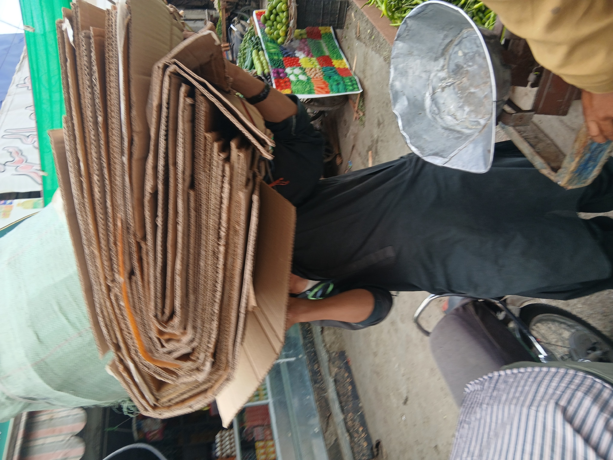A person is on a motorcycle carrying a tall stack of flattened cardboard boxes. The boxes are tan and arranged in a slightly curved stack. The motorcycle has a metal bucket attached and a seat covered in a striped fabric. There is a colorful array of food displayed nearby on a blanket on the ground. The photo was taken in Quetta City Tehsil, Pakistan.