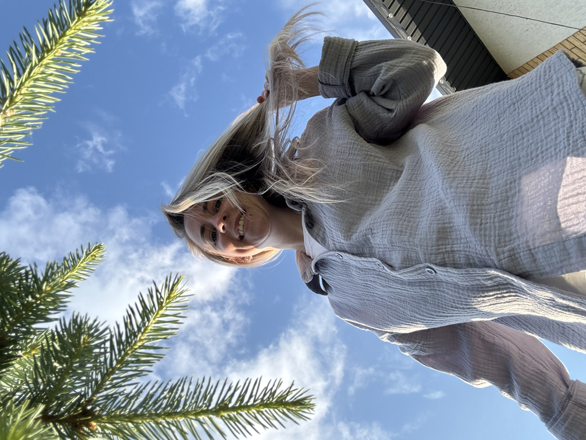 A low-angle outdoor shot captures a blonde woman from the chest up, smiling and holding a lock of her hair. She wears a light-colored, oversized button-up shirt over a white t-shirt. The background is a bright blue sky with scattered white clouds. Pine tree branches are visible in the upper left and right corners of the frame. A portion of a building's roofline is visible in the lower right. The image appears to have been taken in Chaikovsky, Russia.