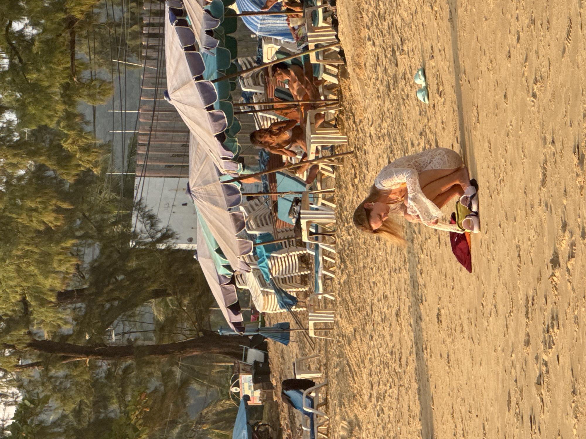 This is a photograph taken on Karon Beach, Thailand, featuring a woman in the foreground kneeling on the sand. She is wearing a white, lacy cover-up over a yellow swimsuit. Her hair is blonde and shoulder-length. In front of her are a red bag and several pairs of shoes.
In the background, there are beach umbrellas providing shade for beach chairs. A couple is seated on chairs under one of the umbrellas. The woman is wearing a white bikini, and the man is wearing orange swim trunks. Further back, stacks of empty beach chairs are visible, as are more umbrellas and a few other beachgoers. A motorbike is parked on the left side of the image, and a signpost is also present. The sand is a light, golden color, and the lighting suggests it is late afternoon or early evening, with the sun casting long shadows. The environment is a tropical beach with palm trees visible in the background, along with some buildings and a fence.