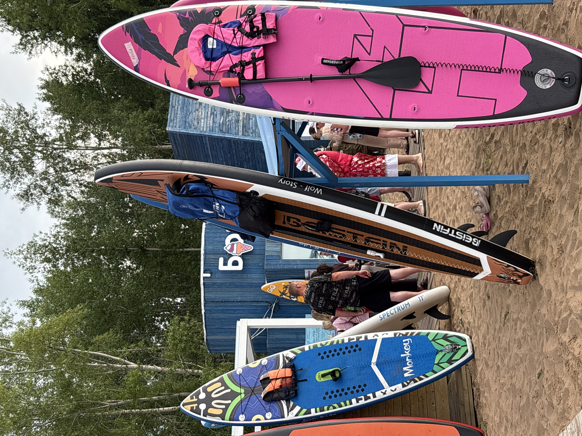 This media file shows a collection of stand-up paddleboards (SUPs) displayed outdoors on a sandy beach, likely in Chaikovsky, Russia. The boards are stacked horizontally and arranged vertically, with some leaning against a blue wooden structure. The overall scene appears to be set during daylight hours, with an overcast sky suggesting a potentially cool or temperate day.
The subjects include several people who are either standing near or interacting with the SUPs. One person in the foreground is wearing a black t-shirt with repeating white text that reads "STYLE READY" and "STYLE". Another person is wearing a red and white patterned garment. The SUPs themselves are prominent objects, featuring various colors and designs. One large pink board has a pink life vest and paddle attached. Another board, dark with brown accents, has the text "Wolf - Story" and "IGEIS ISTFIN" visible. A white board is labeled "SPECTRUM 11", and a blue board has "Monkey" written on it along with a green leaf graphic. A red board is partially visible at the bottom.
The setting is a beach with sand and some trees visible in the background, indicating an outdoor recreational area. The blue wooden building might be a rental shop or a facility related to water sports. The presence of multiple SUPs and people suggests an activity or event related to paddleboarding, such as rentals, a lesson, or a casual gathering. The general atmosphere seems relaxed and focused on the water sports equipment.