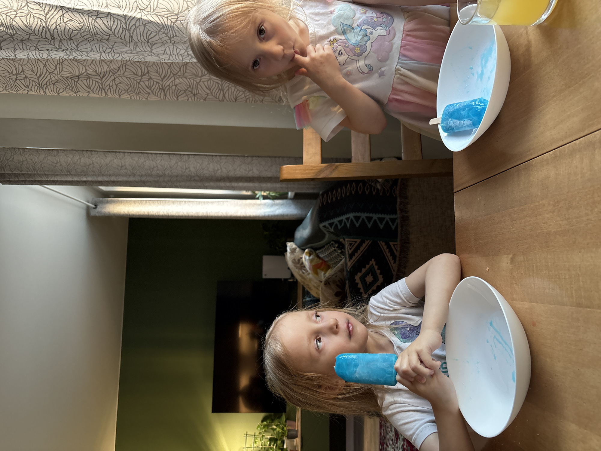 Two young girls are at a wooden table indoors eating blue popsicles in white bowls. One girl is in the upper portion of the image and is seated on a wooden chair. The other girl is in the lower portion of the image. The room has light-colored walls, a green accent wall, patterned curtains, and decorative rugs. The scene is not in Krasnoyarsk, Russia.