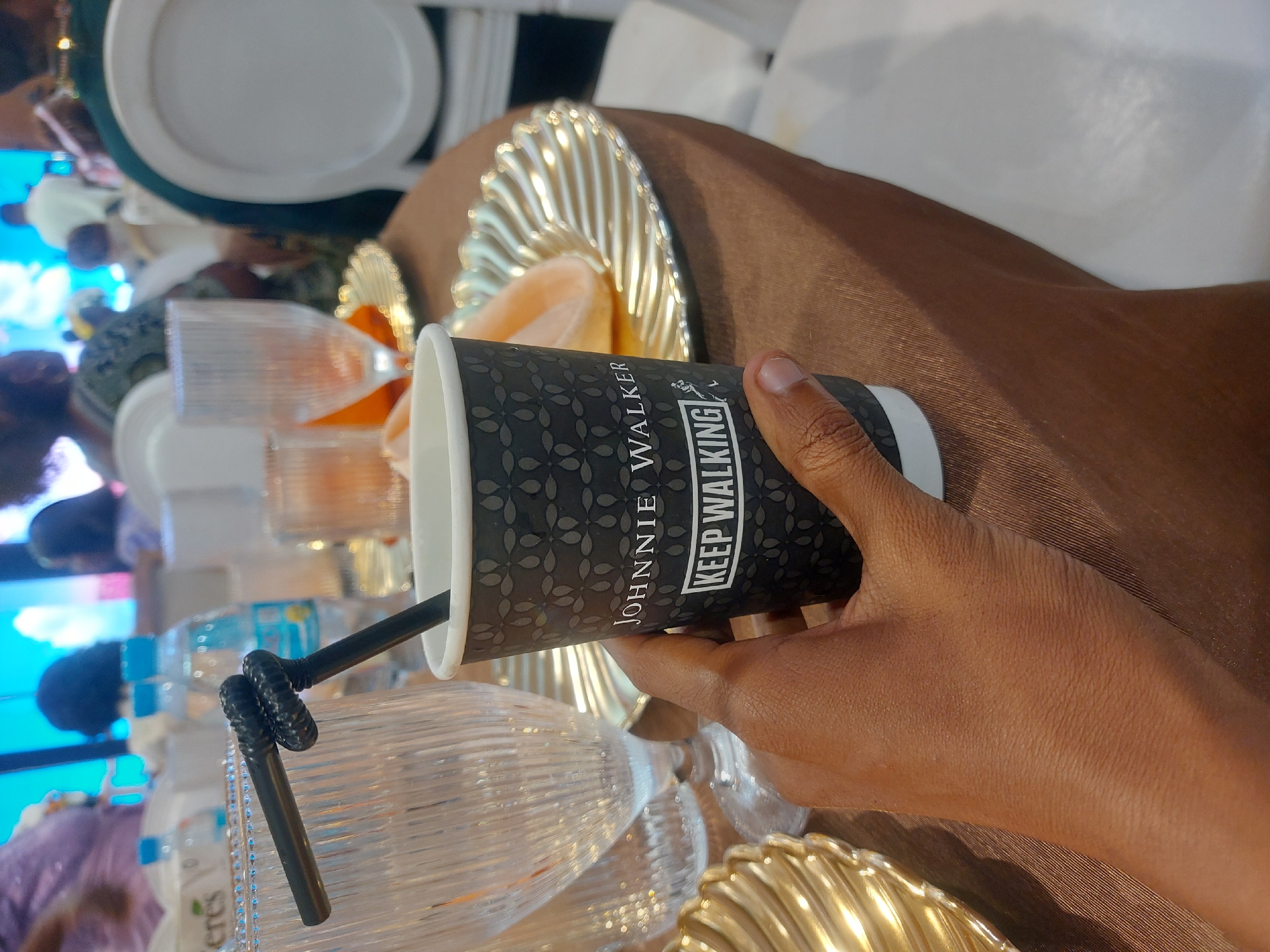 A person holds a "Johnnie Walker Keep Walking" cup with a black straw in a dimly lit event setting. The table is covered with a brown cloth and decorated with golden, ornate plates and transparent glasses. People are visible in the background, seated at tables, suggesting a banquet or a formal gathering. It is possible that this event took place in Dar es-Salaam, Tanzania, but this cannot be confirmed with certainty from the image.