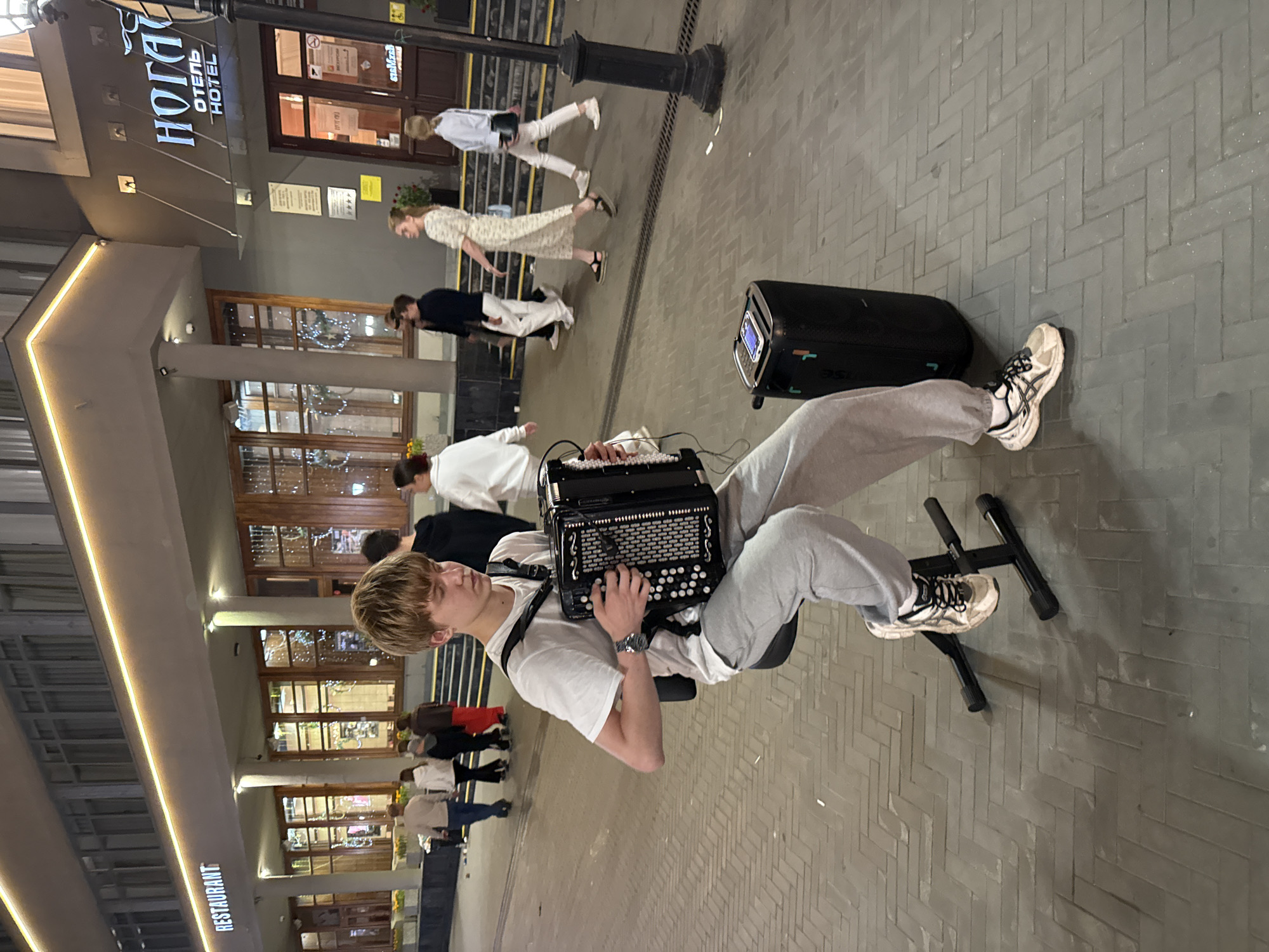 A young male is seated on a black stool on a paved pedestrian walkway, playing a black accordion. He is wearing a white t-shirt, grey sweatpants, and white athletic shoes. To his right is a black portable speaker with a phone resting on top. In the background, a building with a lit "RESTAURANT" sign is visible above a row of large windows. Further down the street, a sign with "НОТА" and "ОТЕЛЬ HOTEL" is illuminated. Several people are walking in the background, some entering or exiting the building. The scene appears to be a public street in Kazan, Russia, likely in the evening.