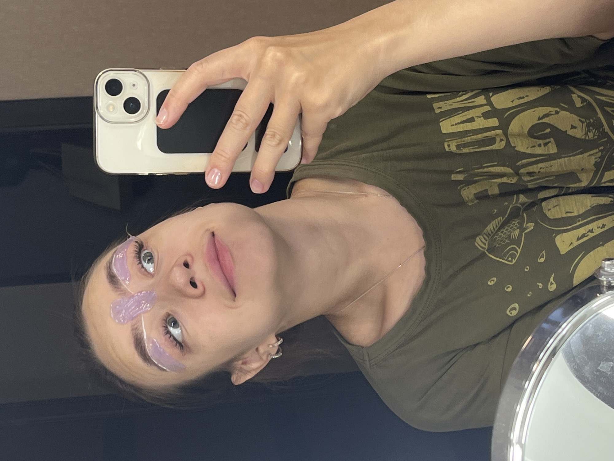 A young woman with light skin and brown hair is taking a selfie in what appears to be a bathroom. She is wearing a dark green t-shirt with a graphic print of a fish and text. Her eyebrows are covered with a purple substance and white strips, suggesting a beauty treatment. She is holding a white smartphone in her right hand, capturing her reflection in a mirror. The background is blurred, showing a wall with a brownish-grey texture and a dark vertical surface. There are no other people or animals visible in the image. The location in Chaikovsky, Russia cannot be determined from the visual information provided.