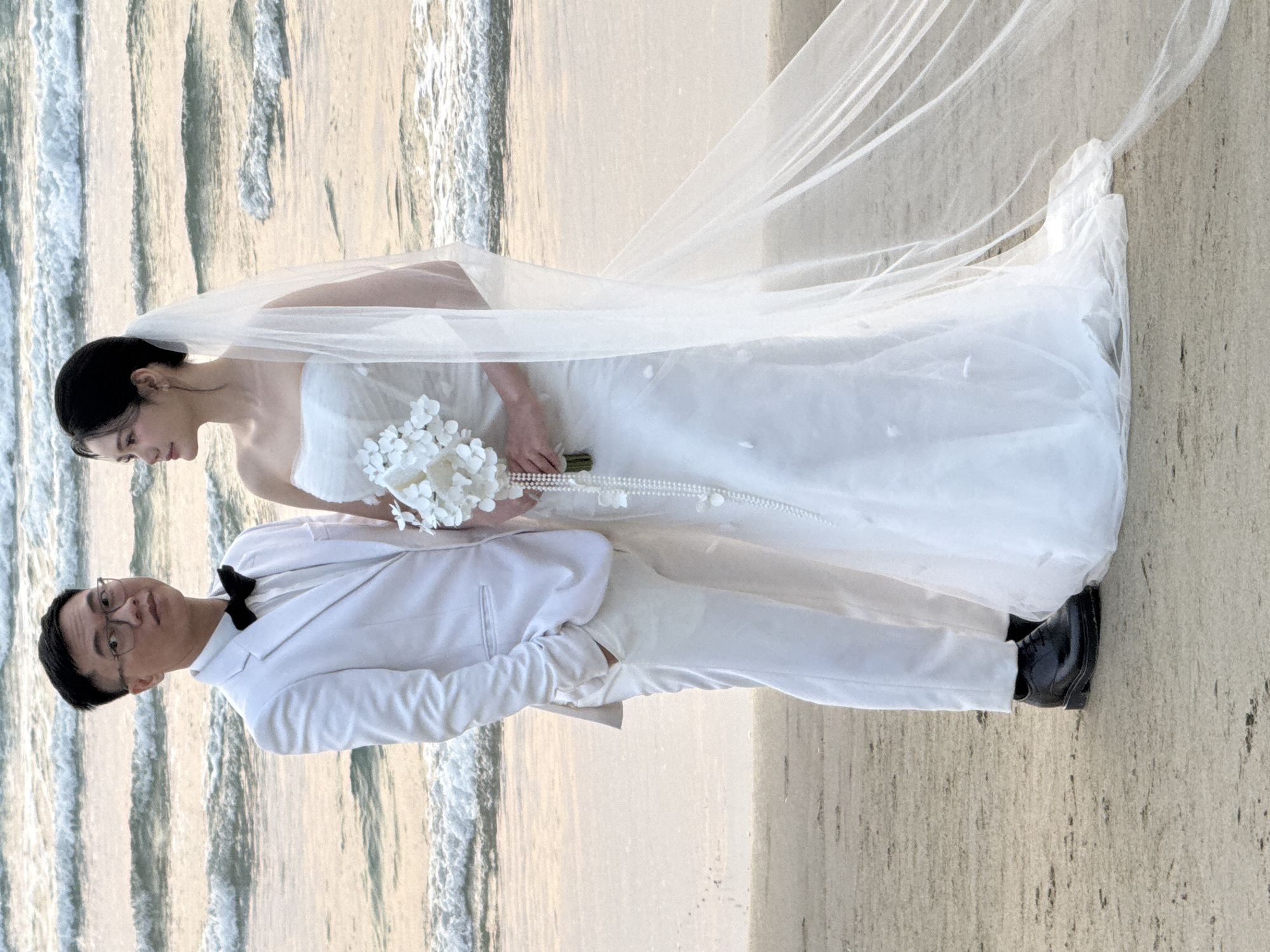 A wedding photo depicts a bride and groom on a sandy beach with waves in the background. The bride wears a white strapless wedding gown and a long veil. She is holding a bouquet of white flowers. The groom is wearing a white suit with a black bow tie and black shoes. The groom stands beside the bride and looks forward. The location is consistent with a beach in Đà Nẵng, Vietnam.