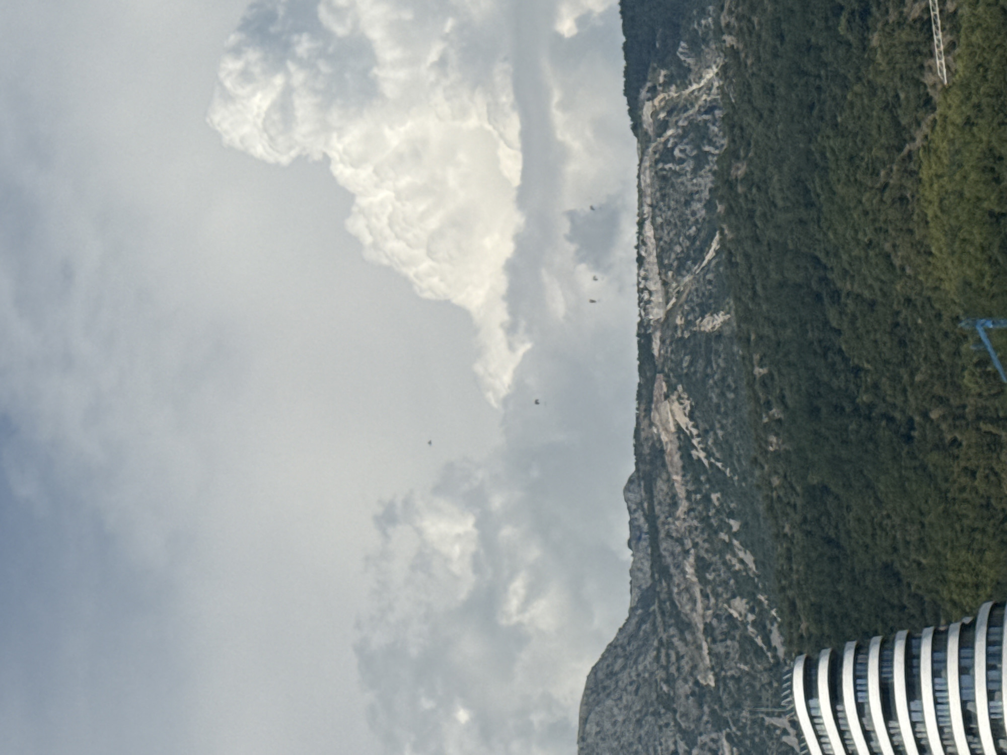 The image captures a dramatic landscape where a steep, heavily forested mountain dominates the right side of the frame, meeting a cloudy sky on the left. Several small, dark figures, possibly birds or paragliders, are visible in the sky, suggesting an outdoor activity is taking place. In the foreground, on the lower right, a modern building with a distinctive ribbed facade is partially visible, hinting at human development integrated into the natural environment. The lighting suggests it could be daytime, with the clouds obscuring direct sunlight. The scene conveys a sense of rugged natural beauty contrasted with contemporary architecture, with the vastness of the sky and mountain evoking a feeling of awe or adventure. The location is identified as Boreti, Montenegro. There is no visible text in the image.