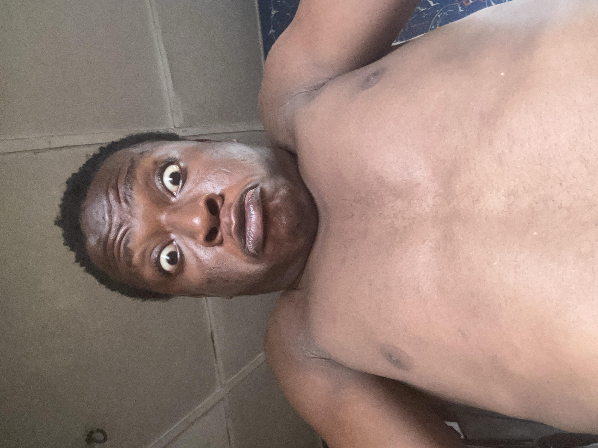 A shirtless person with dark skin is looking directly at the camera with a surprised expression. The background appears to be an indoor space with light-colored walls, but specific details about the location, including the city, are not discernible.