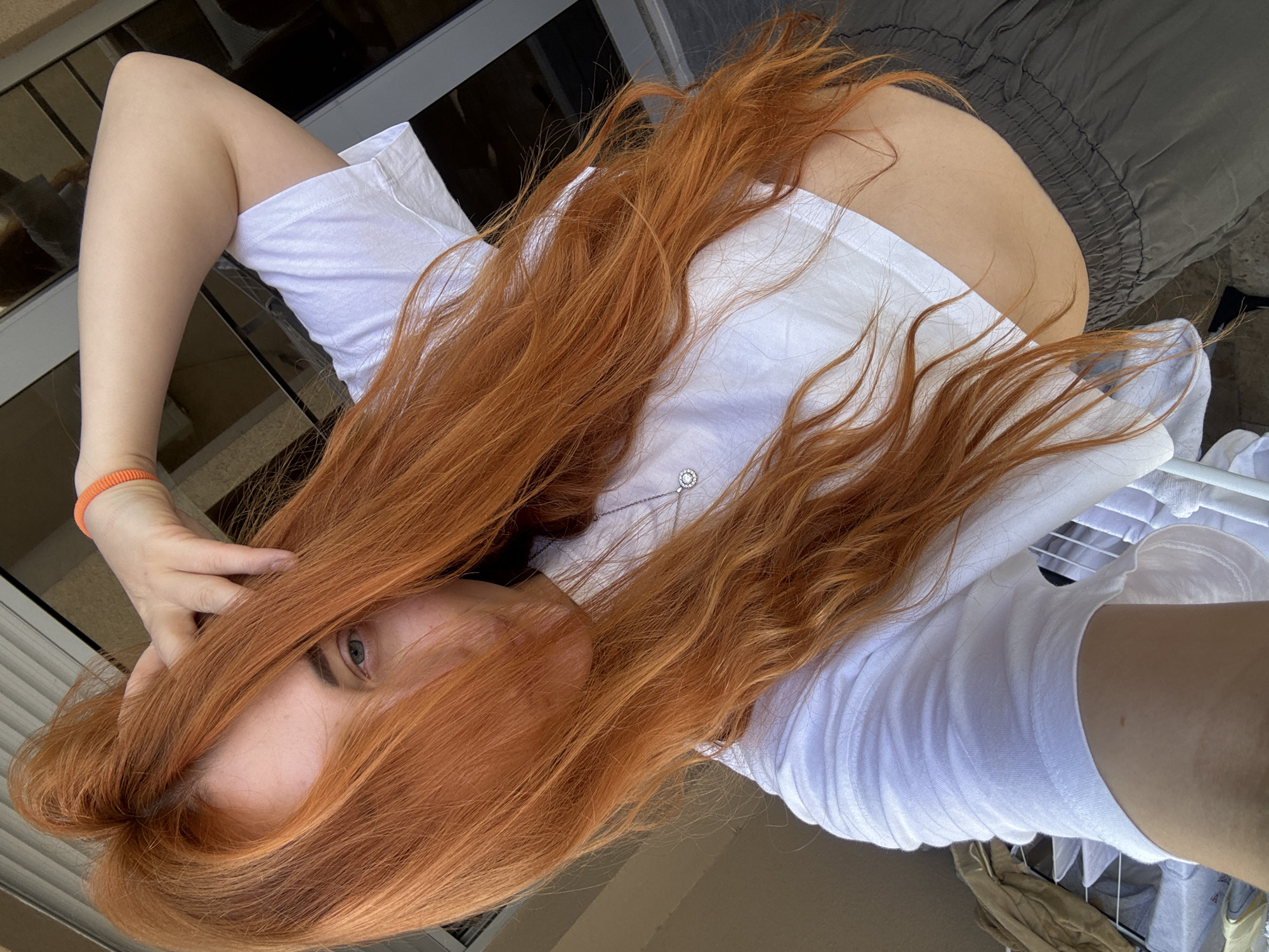 A woman with long, red hair poses for a selfie outdoors. She wears a white t-shirt and gray pants. She has fair skin, and an orange bracelet on her left wrist. Part of her face is visible, including one eye. The background shows an urban setting, possibly in Budva, Montenegro, with a window and a washing rack visible.