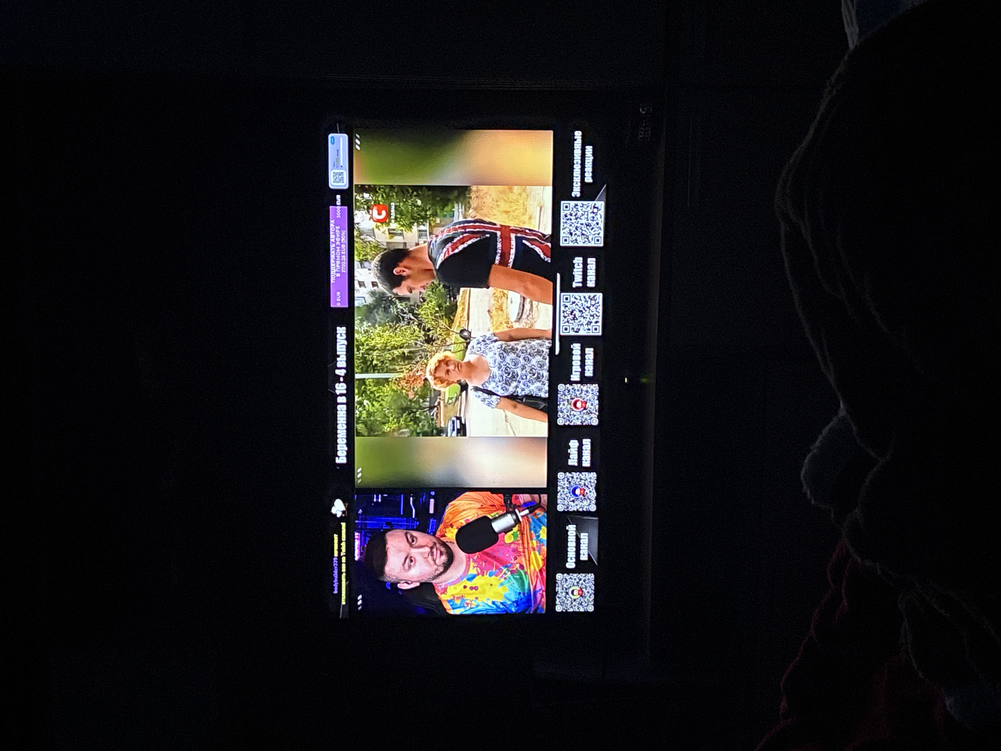 The image shows a television screen displaying a YouTube video in Russian. The video features three segments: a man wearing a Union Jack t-shirt, a woman in a white floral blouse, and a man wearing a brightly colored shirt and using a microphone. QR codes and text in Russian surround the video segments. The text "Беременна в 16-4 выпуск" is visible, which translates to "Pregnant at 16 - episode 4". The screen also shows donation amounts in EUR. The photo was taken in a dark environment. There is no indication that the image was taken in the city of Chornomorskoe, Ukraine.
