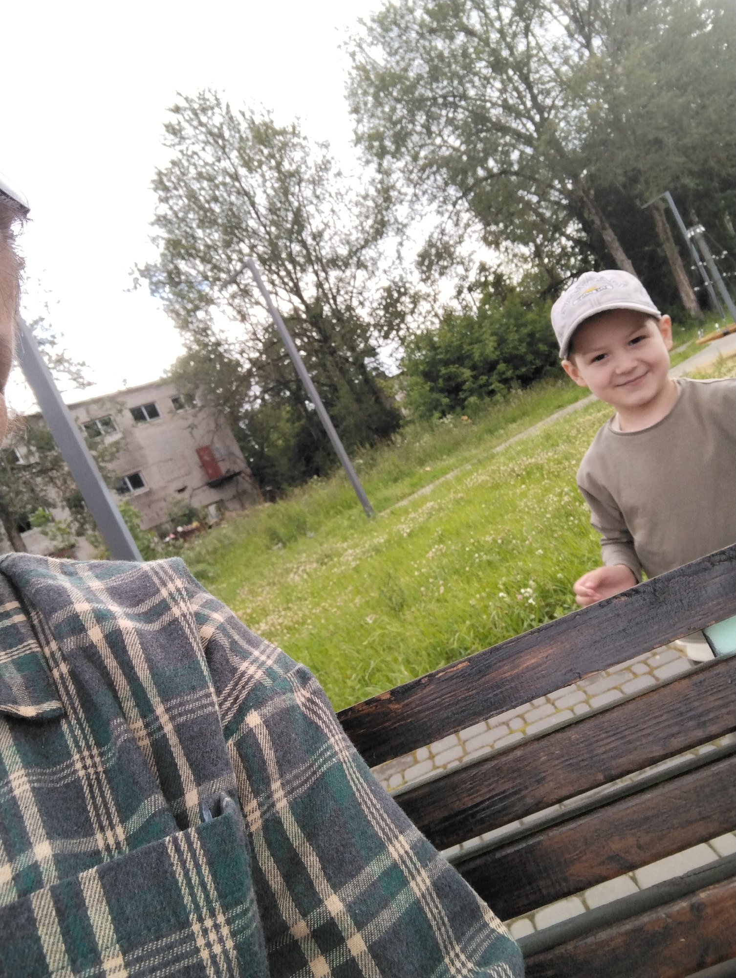 This media file shows a young boy wearing a grey baseball cap and a khaki shirt, smiling at the camera. He is sitting on a wooden bench, with a grassy area and trees in the background. To the left of the frame, a person wearing a green and tan plaid shirt is partially visible, also sitting on the bench. In the background, there is a weathered, multi-story building with several windows. The scene appears to be outdoors, likely during the daytime, with overcast skies. The overall setting suggests a park or an outdoor recreational area. The boy's smile conveys a sense of happiness or amusement. There is no visible text in the image.
