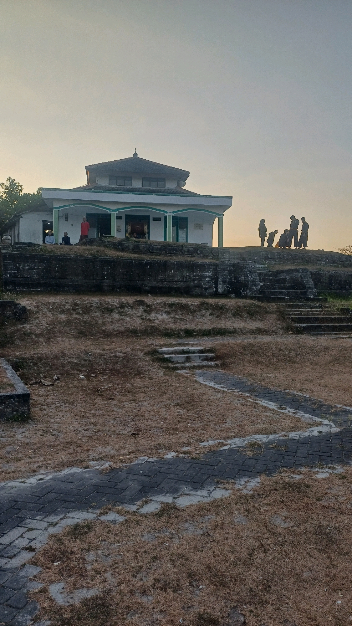 The image depicts a mosque located in Tlogopatut, Indonesia, bathed in the soft light of either dawn or dusk. The mosque, painted white with green trim, stands prominently on a raised platform constructed of stone. A tiled roof crowns the structure. In front of the mosque, a group of approximately five people are visible, some standing and some appearing to be engaged in prayer or religious contemplation.
The foreground of the image is dominated by dry, brown grass and a pathway made of interlocking dark gray bricks with lighter gray grout. This pathway winds its way up towards the mosque, suggesting it's a pedestrian access route. The terrain around the mosque appears somewhat uneven, with terraces and stone retaining walls indicating a sloping landscape. The sky is a pale, hazy hue, consistent with either early morning or late evening light. The overall impression is one of a serene, possibly sacred, outdoor space.