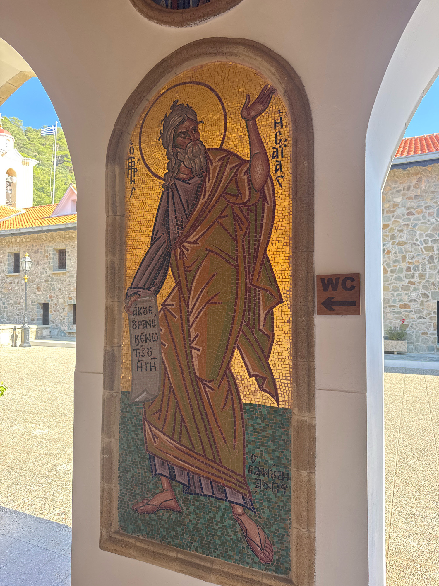 A mosaic depicting a biblical figure is mounted on the arched wall of a building in Tsakistra, Cyprus. The mosaic features a bearded man with long hair, wearing a robe, and holding a scroll. He has his right arm raised. Greek script is visible on the scroll and also vertically on the right side of the mosaic. To the right of the mosaic, a wooden sign with "WC" and an arrow pointing left is affixed to the wall. The building appears to be part of a monastery or church complex, with stone walls and red-tiled roofs visible in the background, along with a bell tower and the Greek flag. The ground in front of the building is paved with light-colored stones. The scene is illuminated by natural sunlight.
