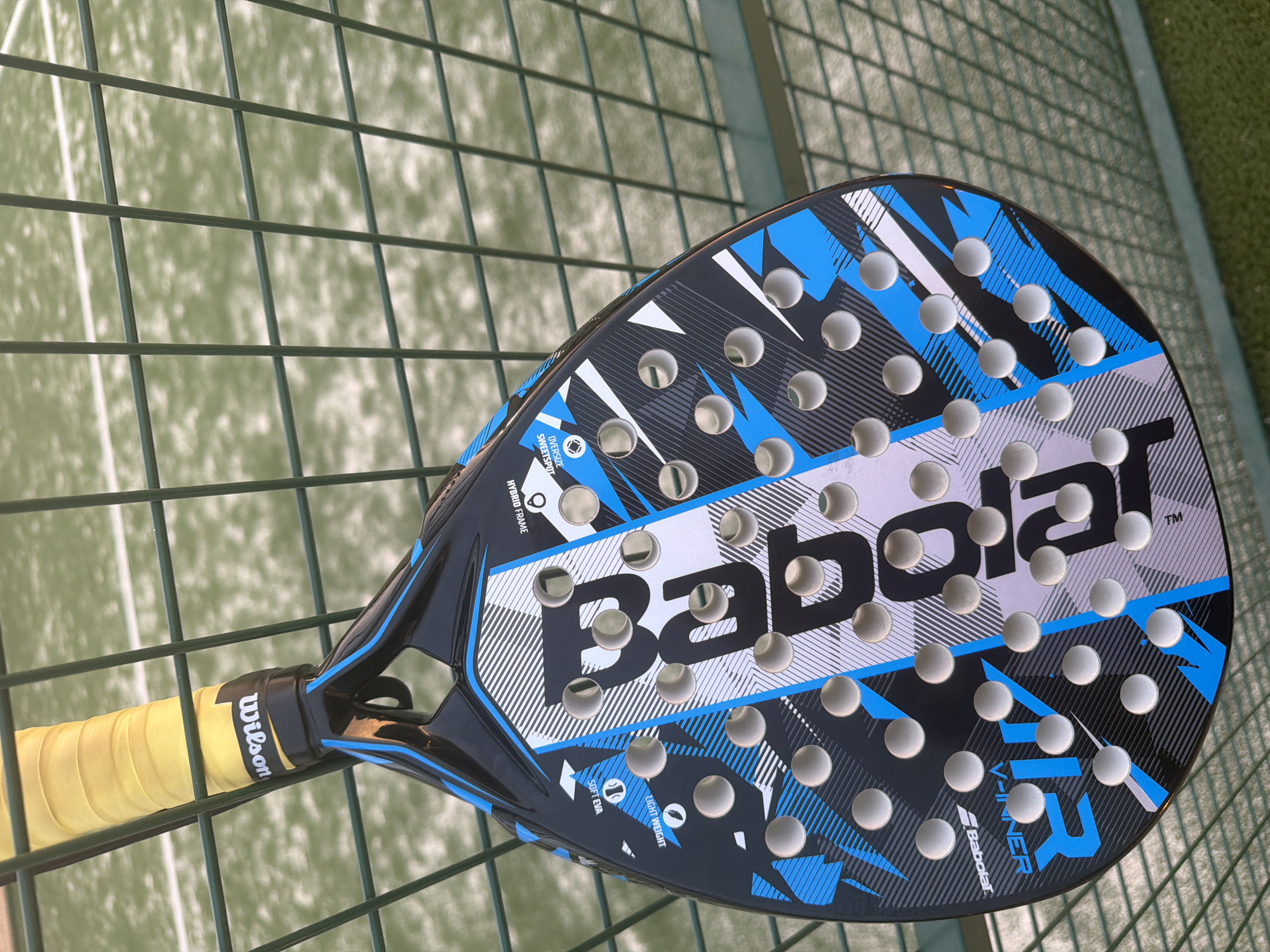 A close-up, top-down view shows a black and blue padel racket with the brand name "Babolat" printed in large white lettering. The racket is resting against a green chain-link fence, with a green, artificial turf padel court visible in the blurred background. The racket has a yellow grip and the "Wilson" logo is visible on the grip. The racket is positioned at an angle, with the head of the racket facing towards the right side of the frame. The image appears to have been taken outdoors, likely in the city of Finestrat, Spain, given the distinctive green artificial turf courts commonly found in the region and the brand of racket, popular in Spain. No people are visible in the image.