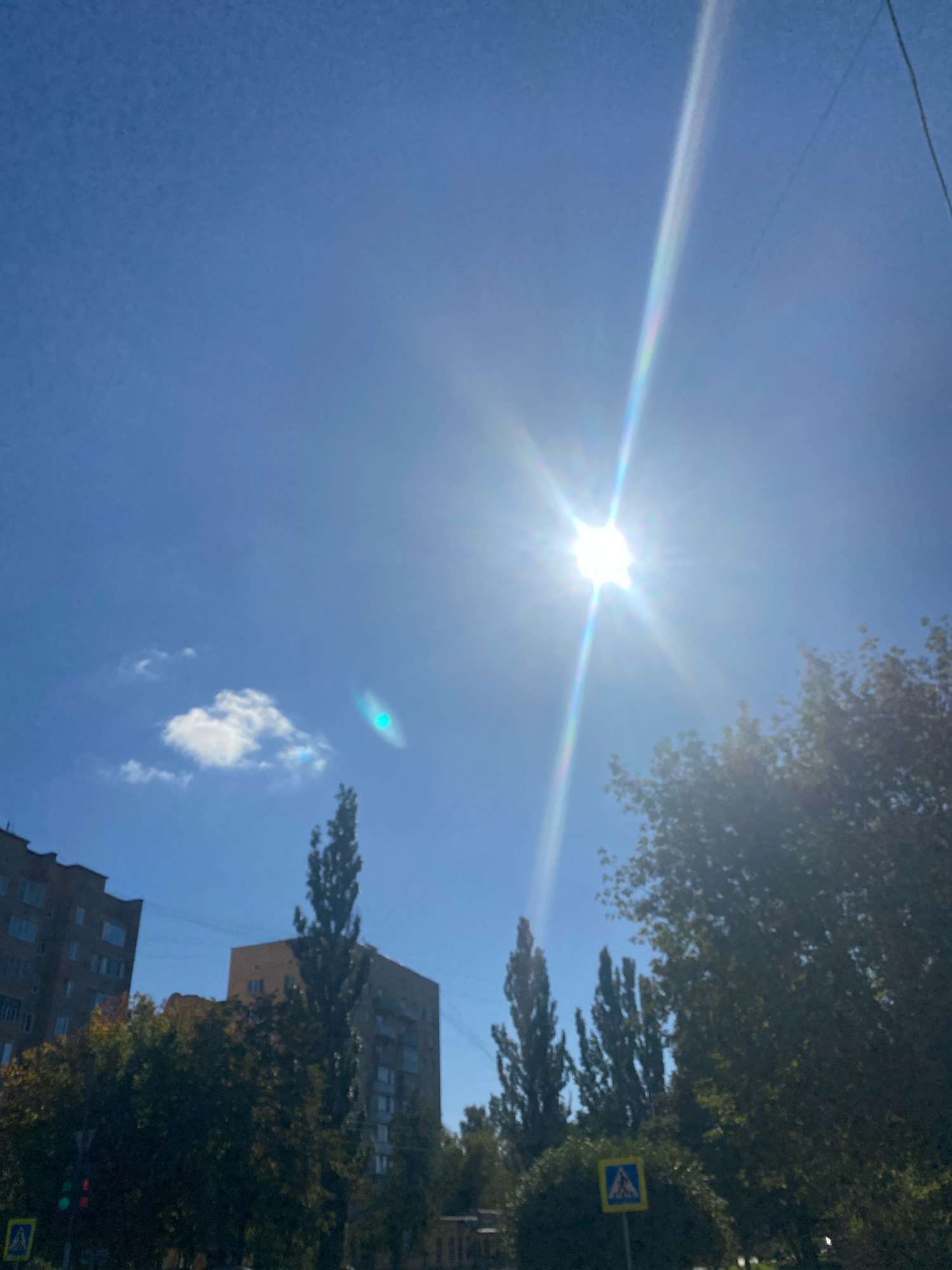 The image shows a bright, sunny day with the sun prominently featured in the upper center, casting a lens flare and rays across the blue sky. A single, small, white cloud is visible to the left of the sun. In the foreground, there are trees with green foliage and a few tall, slender trees. Several multi-story buildings are partially visible behind the trees, suggesting an urban or suburban setting. A traffic light with a green light illuminated is on the left side of the image. A pedestrian crossing sign with a yellow background and a blue symbol indicating a pedestrian crossing is also visible near the bottom left. The presence of buildings, traffic signals, and pedestrian signs indicates this scene is in a populated area, likely a city. Given the prompt's context, this scene is located in Ivanteyevka, Russia. There are no people clearly visible in the image. The overall environment is bright and appears to be daytime.