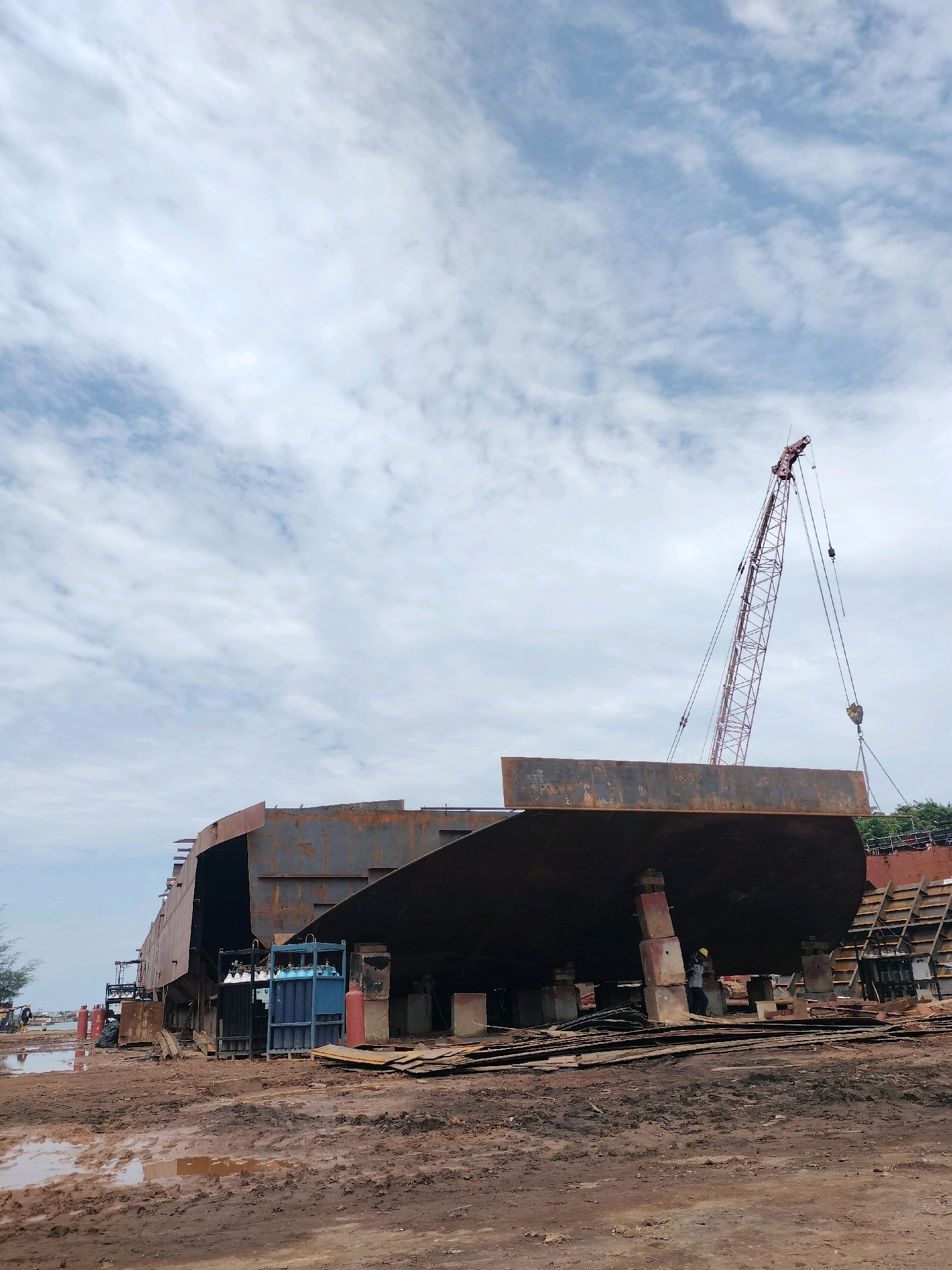 The image shows a shipyard in Batam, Indonesia, with a large ship hull under construction. A red crane is positioned over the hull, with its boom extended and cables lowered, suggesting it is being used to lift or position heavy components. The ship's hull is made of rusted metal, indicating it is in an early stage of construction. The surrounding environment is a muddy yard, with piles of scrap metal and wood scattered around. Several industrial gas cylinders, including blue and black ones, are stacked near the hull. In the distance, there are trees and other structures associated with a shipyard. The sky is cloudy, with a mix of white and blue. The scene depicts industrial activity in a maritime setting.