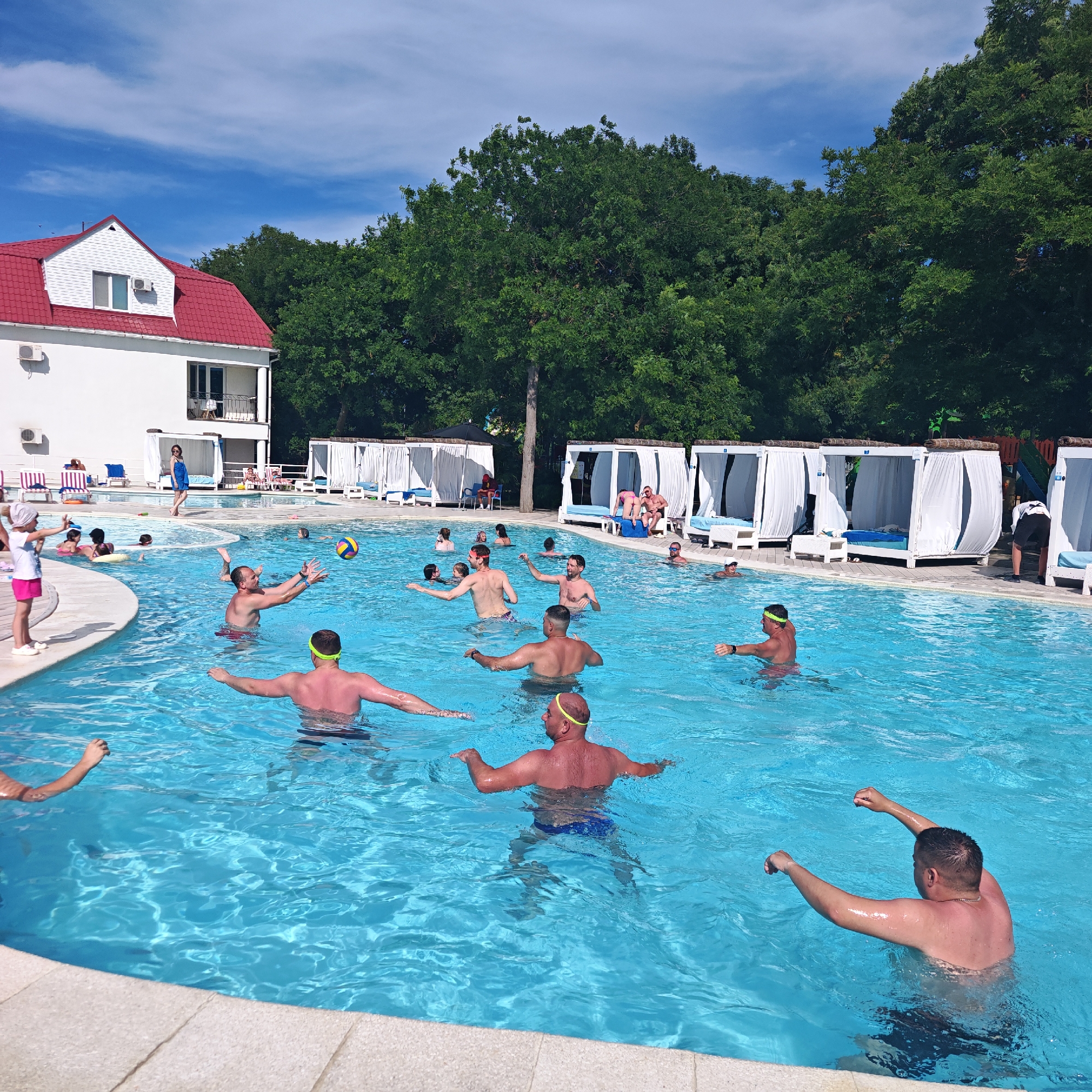 This image depicts a lively scene at an outdoor swimming pool, possibly part of a resort or recreation complex in the vicinity of the city of Shchyolkino, Ukraine. The pool is filled with clear blue water, and a group of people, predominantly adults, are engaged in various water activities. In the foreground, several men are visible, some with swim goggles on, appearing to play a game of water volleyball. One man in the mid-ground is reaching up to receive the volleyball, while another in the foreground has his arms outstretched. The pool area is surrounded by lush green trees, and in the background, there is a white building with a red roof, resembling a hotel or administrative building, and several white cabanas with blue cushions. The sky is bright blue with scattered white clouds, indicating a sunny day. A young girl in a pink skirt and white top is standing on the edge of the pool, facing away from the camera. The overall atmosphere suggests a recreational and social gathering.
