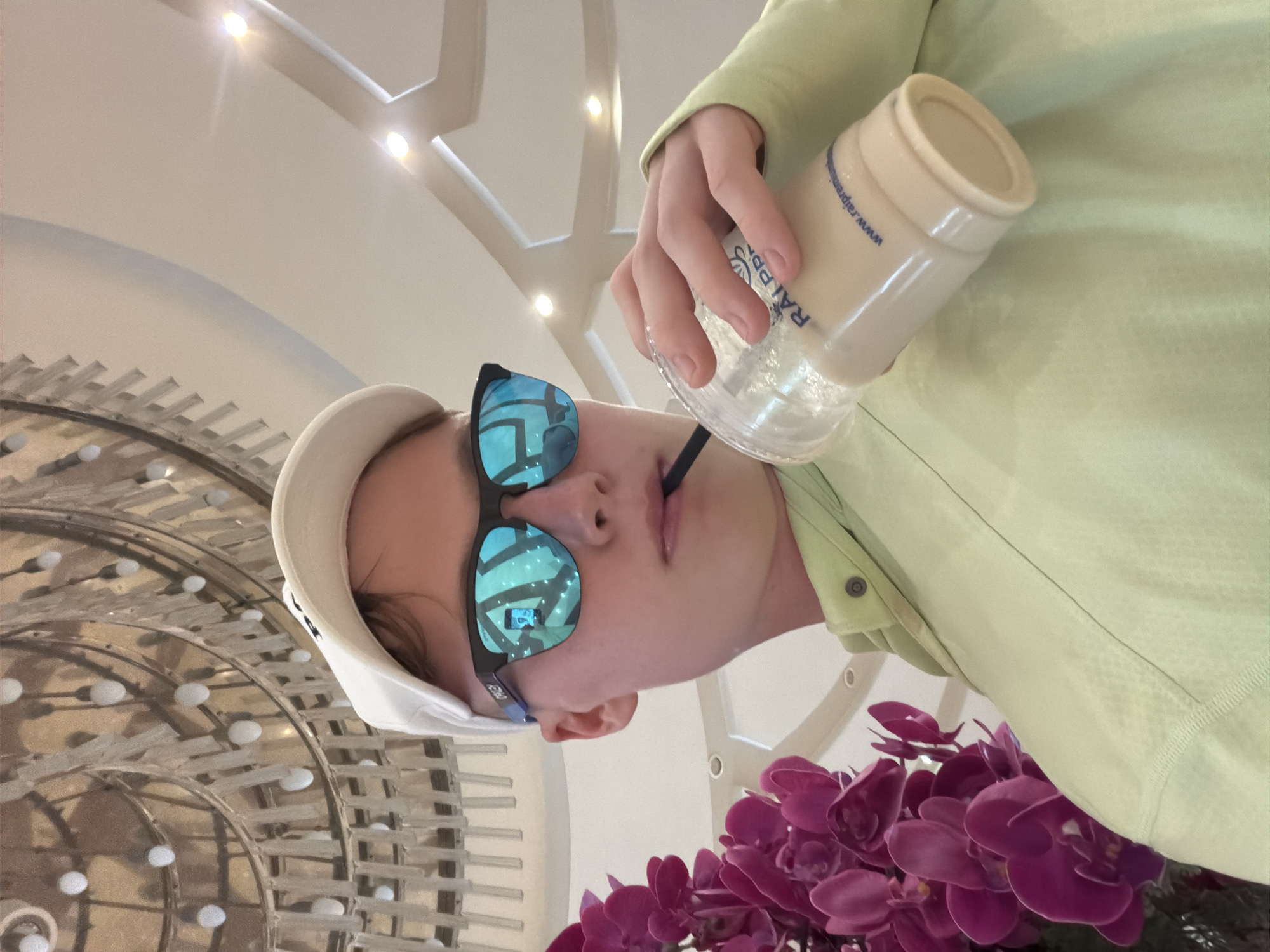 A young person wearing a white baseball cap and blue mirrored sunglasses is taking a sip from a clear plastic cup filled with a light-colored beverage. The person is also wearing a light green long-sleeved shirt. In the background, a large, ornate chandelier hangs from the ceiling, and a cluster of vibrant purple orchids is visible on the left side of the frame. The person's face is partially obscured by the sunglasses and the angle of the shot. The environment appears to be indoors, possibly a hotel lobby or restaurant, given the presence of the chandelier and elaborate decor. The person's clothing and accessories suggest a casual yet stylish presentation. The reflective sunglasses show a distorted image of the surroundings, including lights and possibly a phone. The cup has blue lettering that reads "RAI PREMIUM" and a website address. The overall scene conveys a sense of relaxed enjoyment. The image is set in the city of Kemer, Turkey.