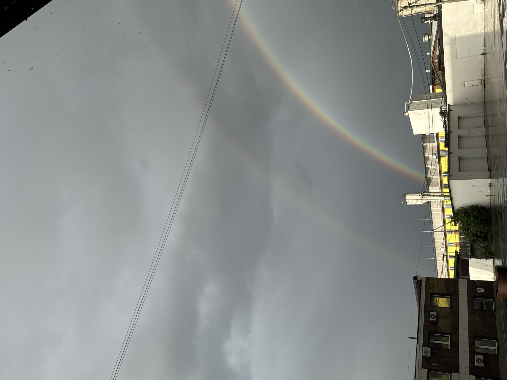 A low-angle outdoor photograph shows a double rainbow in a grey sky over a cityscape. The sky is overcast with dark, heavy clouds. The rainbow arcs from the upper left to the upper right of the frame. Buildings are visible on the right side of the image, including a white building with what appears to be loading dock doors and a brown building with multiple windows and air conditioning units. Wires stretch across the sky. The photograph may have been taken after or during rainfall due to the wet appearance of the buildings. The location may be Perm, Russia, as specified in the prompt.