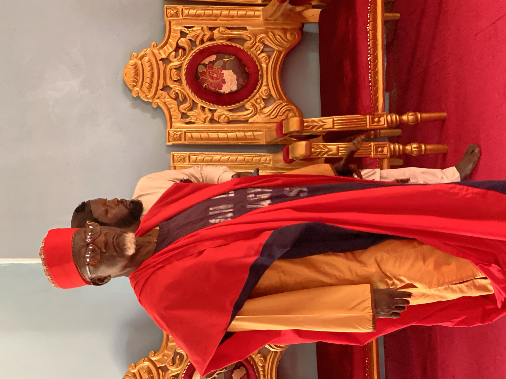 An elder Black man sits on an ornate golden throne, adorned in a red cap, glasses, and a red and blue robe. A second Black man with a beard stands behind him, slightly out of focus. Both men are in Sabon Tasha, Nigeria. The throne sits atop a red carpet against a light blue wall.