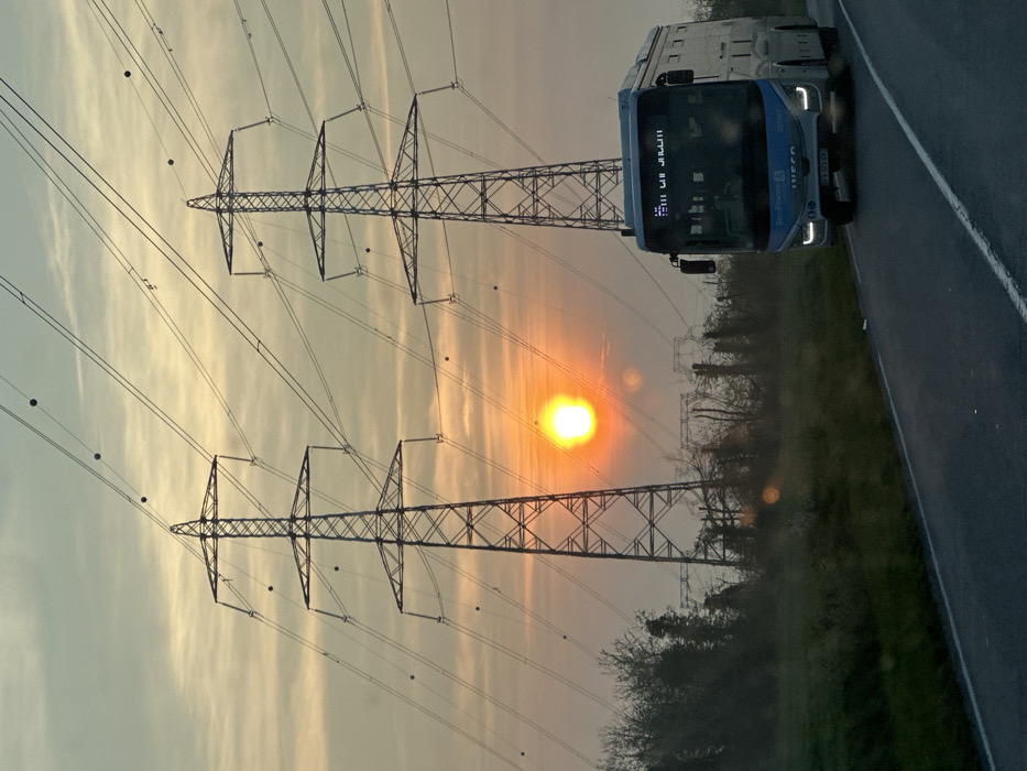 The scene depicts a semi-rural landscape in Marcoussis, France, at sunrise or sunset. A bright, diffused orange-yellow sun is positioned low in the sky, slightly to the left of the image center, casting a warm hue across a pale orange and blue-grey sky with light cloud cover. Two prominent lattice-style high-voltage transmission towers stand against the sky, with numerous power lines extending across the frame. Several dark spherical aviation warning markers are attached to these power lines.
In the foreground, an asphalt road curves from the lower right towards the middle left. A light blue and white Île-de-France Mobilités transit bus, an IVECO model with license plate FB-103-GV, occupies the right side of the road. Its headlights are illuminated, and a digital destination sign on the front displays partially legible text. The bus appears to be in motion, approaching the viewer.
To the left of the road, a strip of green grass is visible, beyond which a dense line of bare deciduous trees and bushes forms a horizon. Further in the background, additional, less distinct power line structures or smaller pylons are visible. No individuals are distinctly present in the visible portion of the image.