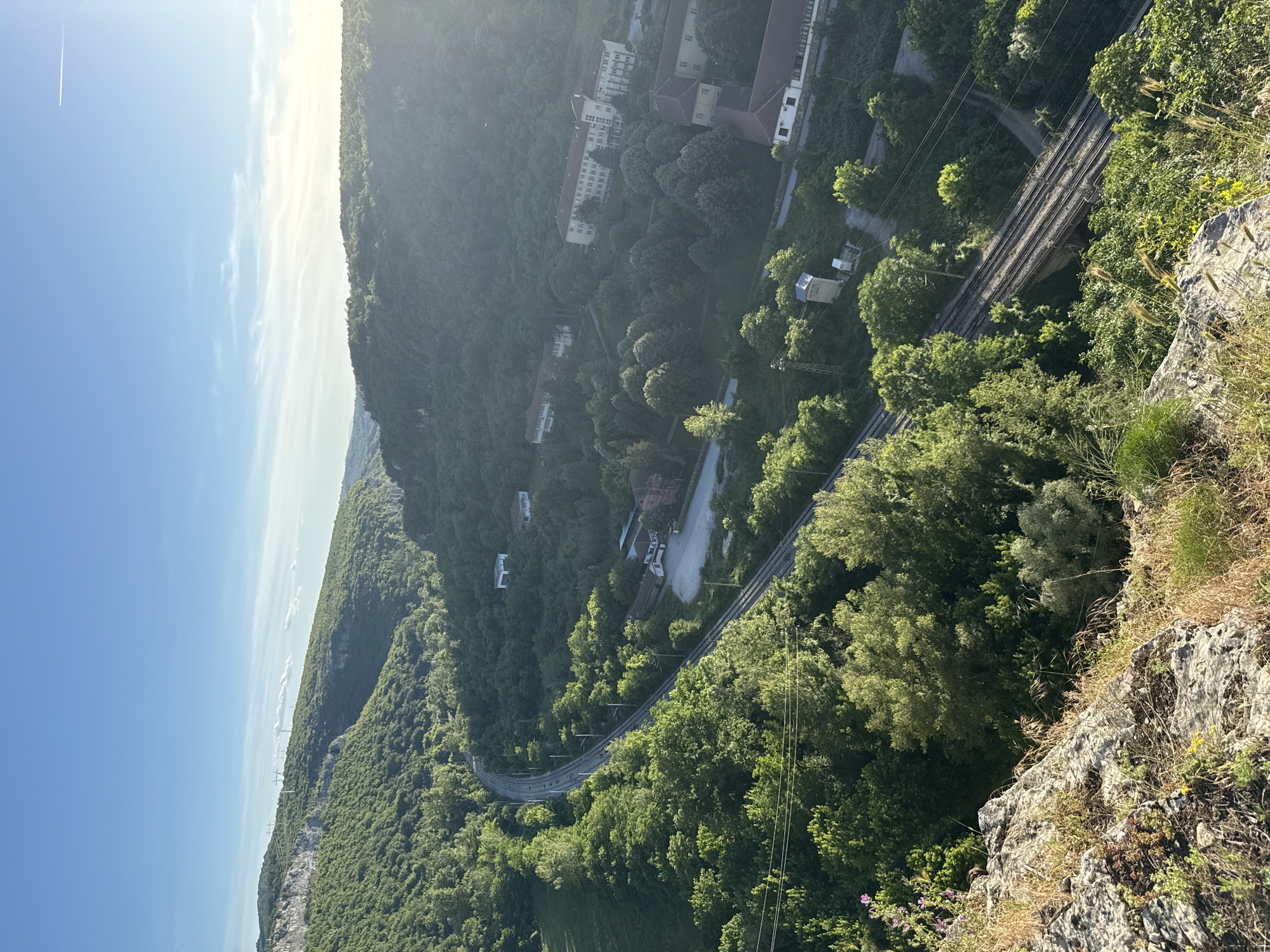 This is a high-angle, medium-shot, vertical photograph taken from a rocky, grassy overlook, capturing a valley with lush green trees, a curving asphalt road, and railway tracks. In the background, several buildings, some appearing to be institutional or residential complexes, are nestled among the trees on the side of the valley. A body of water is not visible in the frame. The sky is bright blue with a few wispy clouds, and the sun is shining from the right, creating lens flare and highlighting the edges of the trees. The overall scene depicts a natural landscape integrated with man-made infrastructure in a valley setting, likely in or near Karlukovo, Bulgaria, given the context.