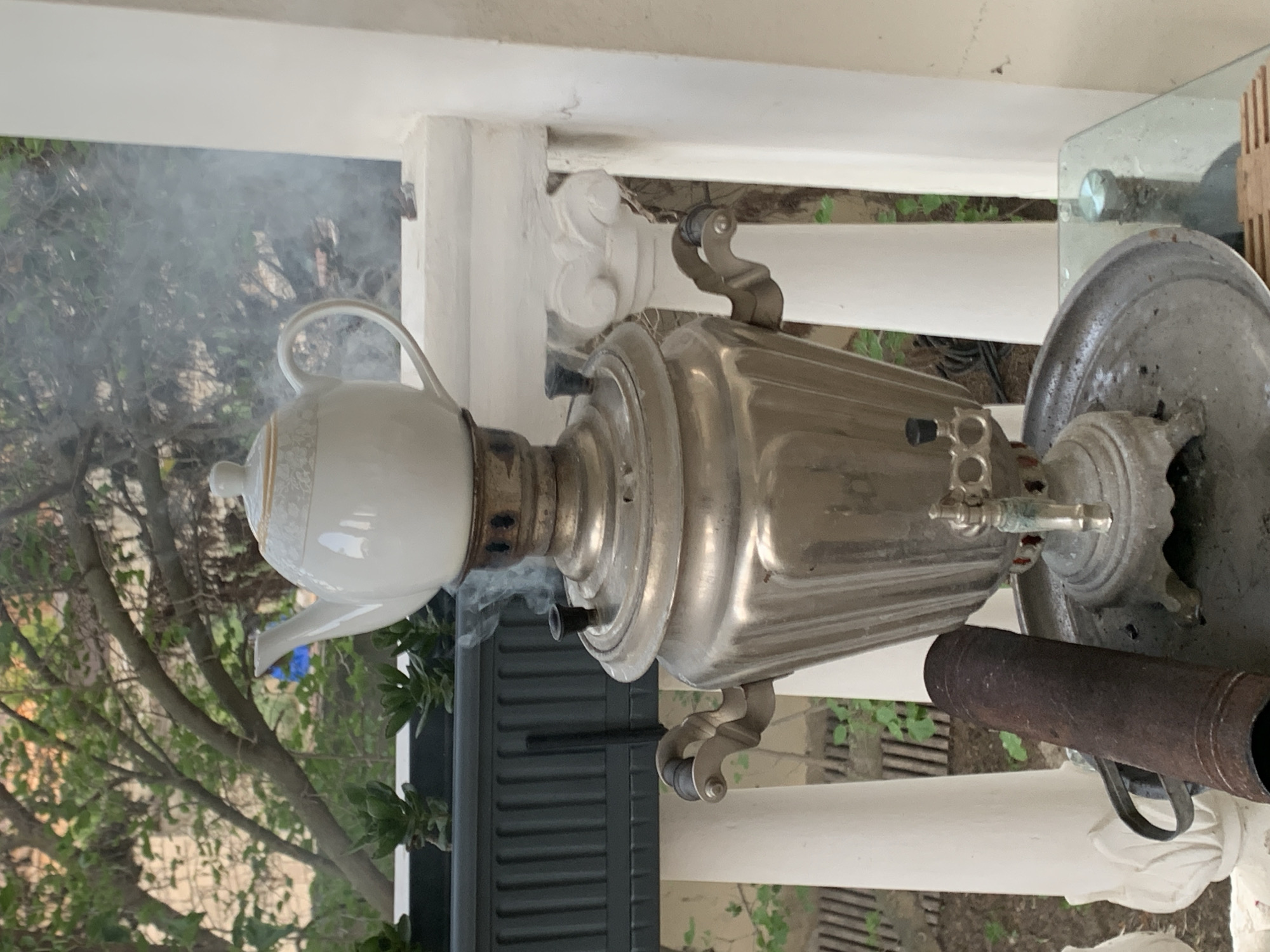 A samovar with a white teapot on top is steaming, suggesting it is in use. The samovar is made of metal and has decorative handles. It sits on a tray with a rusty pipe next to it. In the background, there are white columns, railing, and some greenery. The scene appears to be outdoors, possibly a patio or balcony, with trees and plants visible. The overall impression is one of a traditional setting, likely in a place where samovars are commonly used, such as Khazar Raion, Azerbaijan.