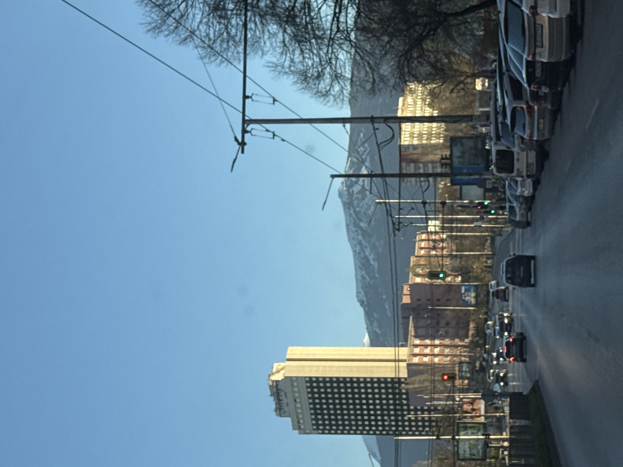 The image depicts a street scene in Sofia, Bulgaria, with a prominent view of the Rhodope Mountains in the background. The camera is likely positioned inside a vehicle on a roadway, looking toward buildings in the downtown area. The main building is a modern, high-rise building with a grid-like façade. A line of cars is seen parked on the right side of the road. A street with traffic lights is visible ahead. Overhead electrical cables and utility poles line the street. The sky is clear and blue.