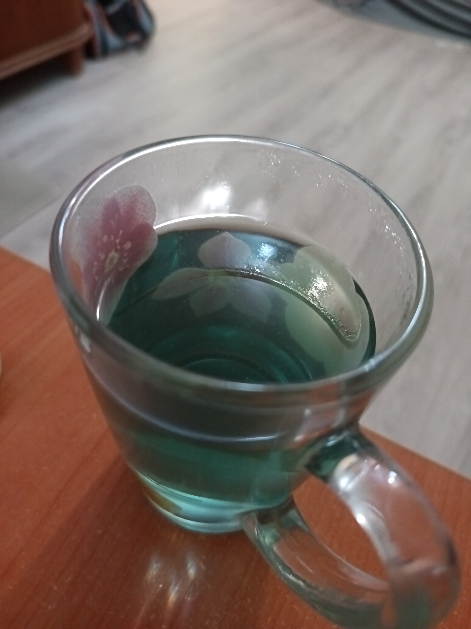 A clear glass mug filled with a greenish-blue liquid sits on a wooden surface. The mug has a floral pattern etched on its exterior, with a pinkish flower visible on the left side and a lighter flower or design on the right. The liquid appears to be tea or a similar beverage. The background is out of focus but suggests an indoor setting with a light-colored floor and a dark wooden piece of furniture. There are no people or animals visible in the image. The scene does not provide any information about the location in the city of Minsk, Belarus.