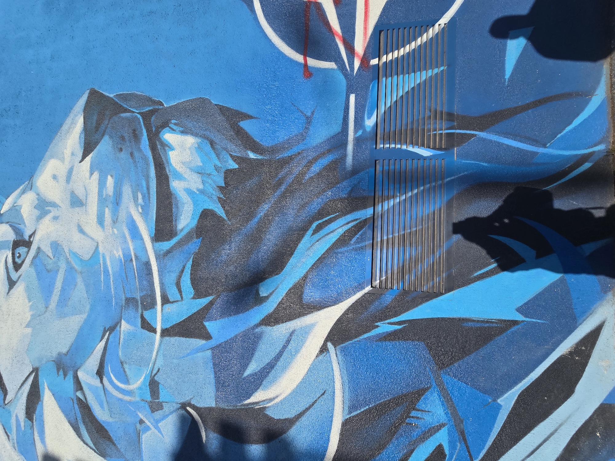 This media file displays a vibrant graffiti mural predominantly in various shades of blue, black, and white, painted on a textured wall. The central subject on the left side is a highly stylized and angular depiction of an animal's face, possibly a tiger or a lion, characterized by sharp geometric forms, flowing lines, and a distinct eye on the far left. The artwork combines strong outlines in black with lighter blue, white, and darker blue fills, creating a dynamic and fierce impression.
Integrated into the right side of the mural is a dark blue rectangular vent or grate with vertical slats, which is partially obscured by the painted elements. Above and to the left of the vent, abstract red and white lines or shapes are visible against the blue background, possibly indicating another layer of graffiti or a tag.
The scene is outdoors, likely during daytime, as indicated by the bright sunlight and the distinct, dark shadows cast across the lower and right portions of the image. These shadows suggest an object or person is standing in front of the wall, possibly the photographer. The wall itself appears to have a slightly rough, granular texture beneath the paint. No explicit text is visible in the artwork.