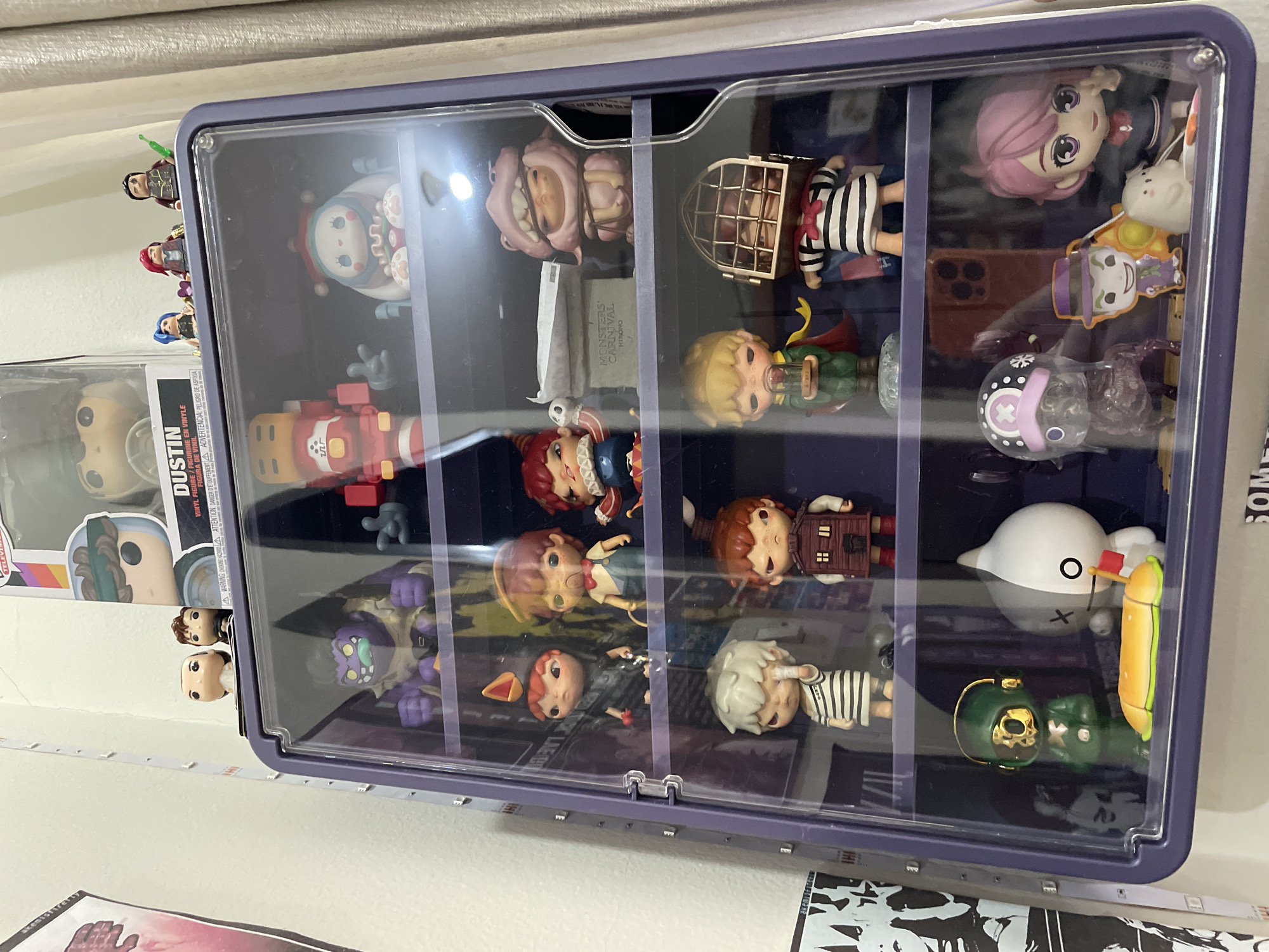 The image displays a collection of collectible figurines organized within a clear, purple-trimmed display case. The case is situated against a plain white wall, with a curtain visible on the right side. The figurines are arranged on multiple shelves, showcasing a variety of characters and styles. Some prominent figures include a red and yellow robot-like character on the top shelf, a purple monster with a green mouth on the second shelf, and several smaller figures with distinct facial features and clothing on subsequent shelves. In the bottom section of the display, a green helmeted figure stands next to a white character with a black circle for an eye. The top of the display case also features additional figurines, including what appears to be a Funko Pop box with a character named "DUSTIN" on it. The image does not provide any information or visual cues that would allow for the determination of its location in Denpasar, Indonesia, or any specific city.